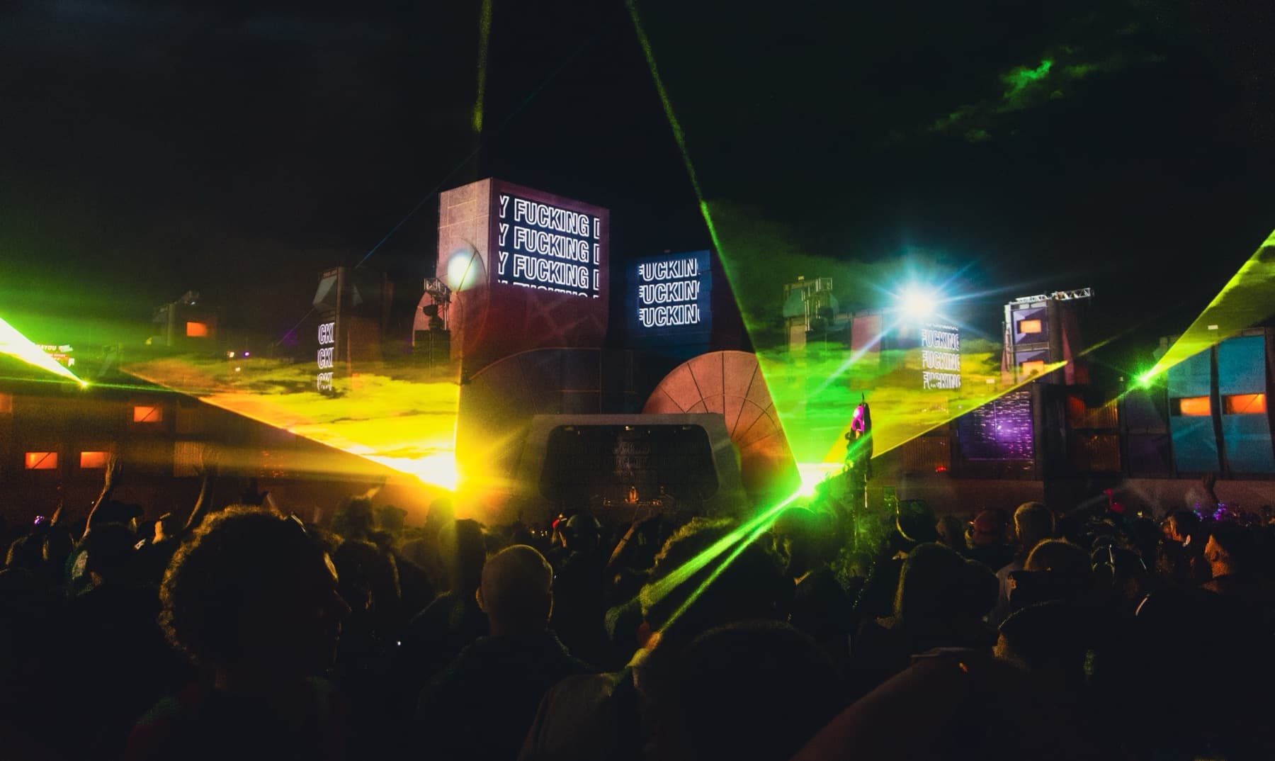 Green and yellow lasers cutting across the night sky over a silhouetted crowd with LED screens on stage