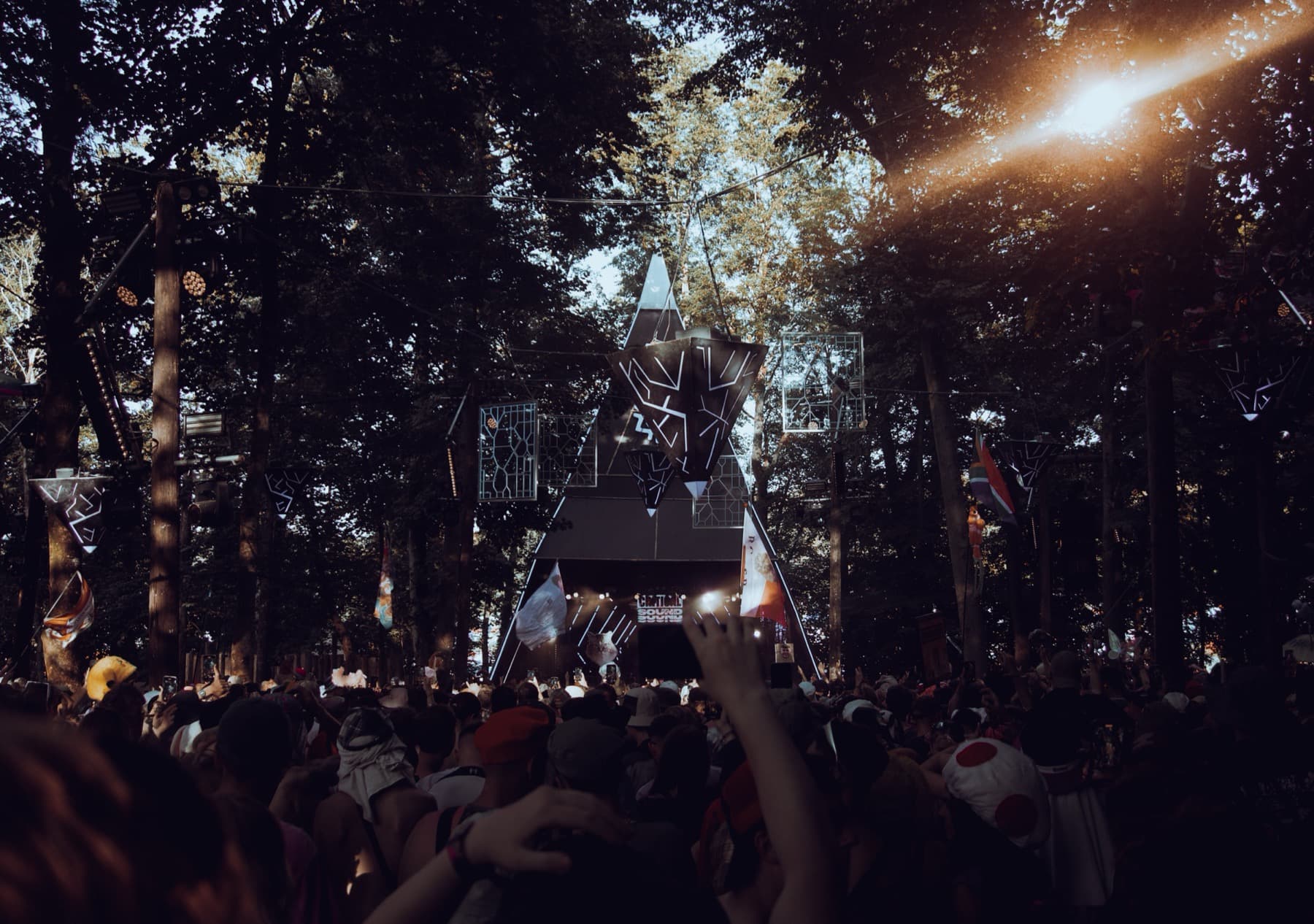 Festival crowd dancing beneath a geometric star-shaped stage structure in a sunlit forest clearing