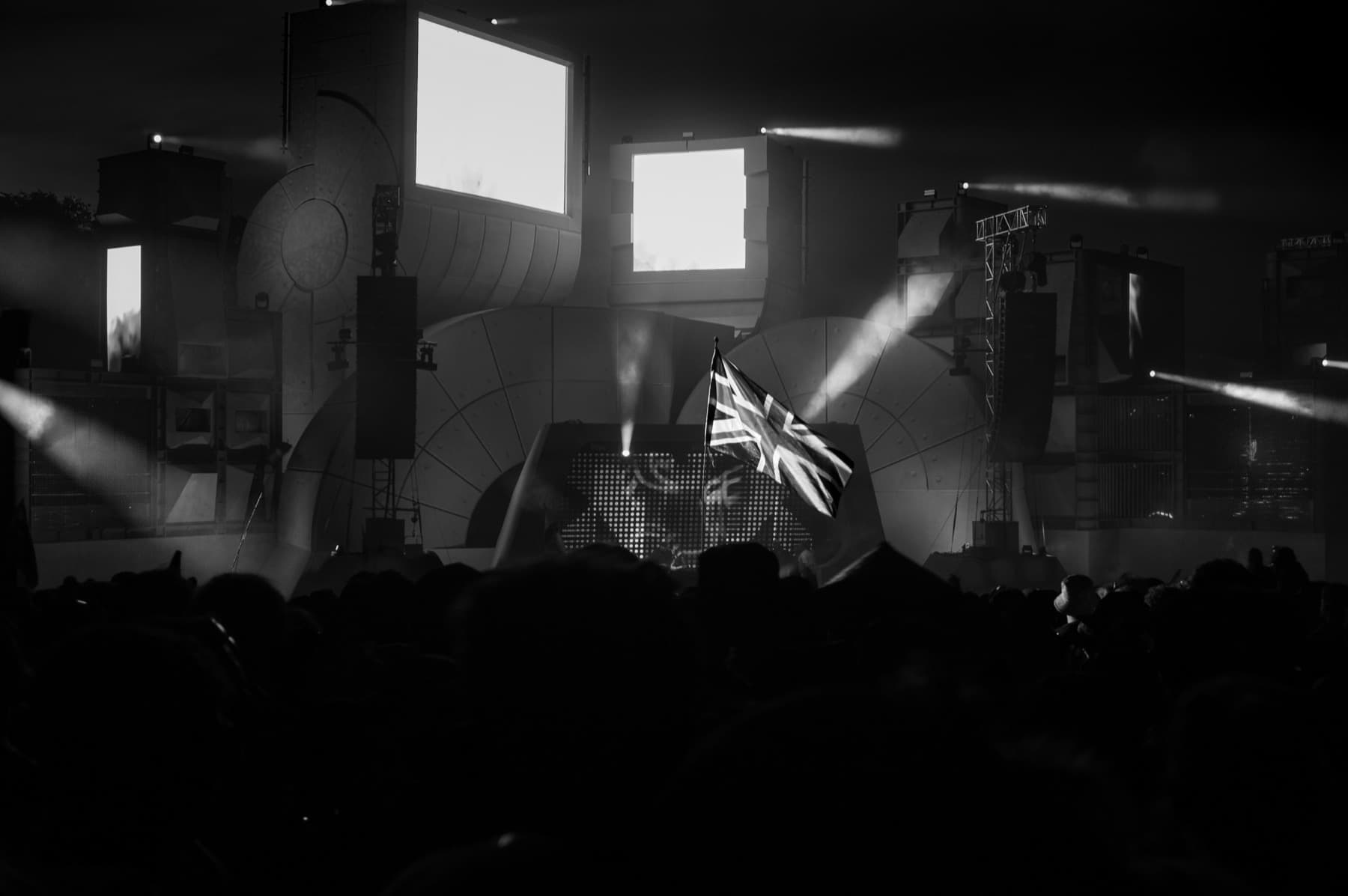 Black and white shot of a Union Jack flag waving in front of a massive industrial stage set with sweeping spotlights