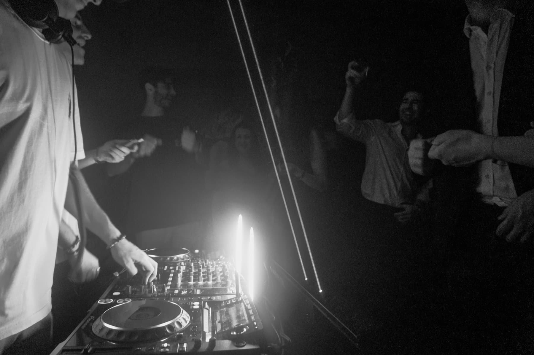DJ mixing on CDJs with hands raised from the crowd visible in dramatic backlight and haze