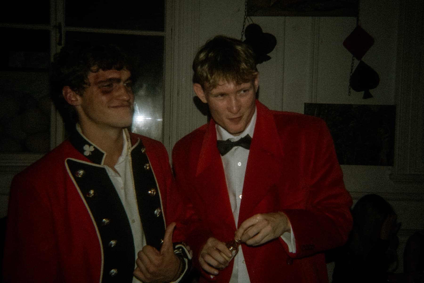 Two friends in red military-style jackets and bow ties with playing card decorations hanging behind them