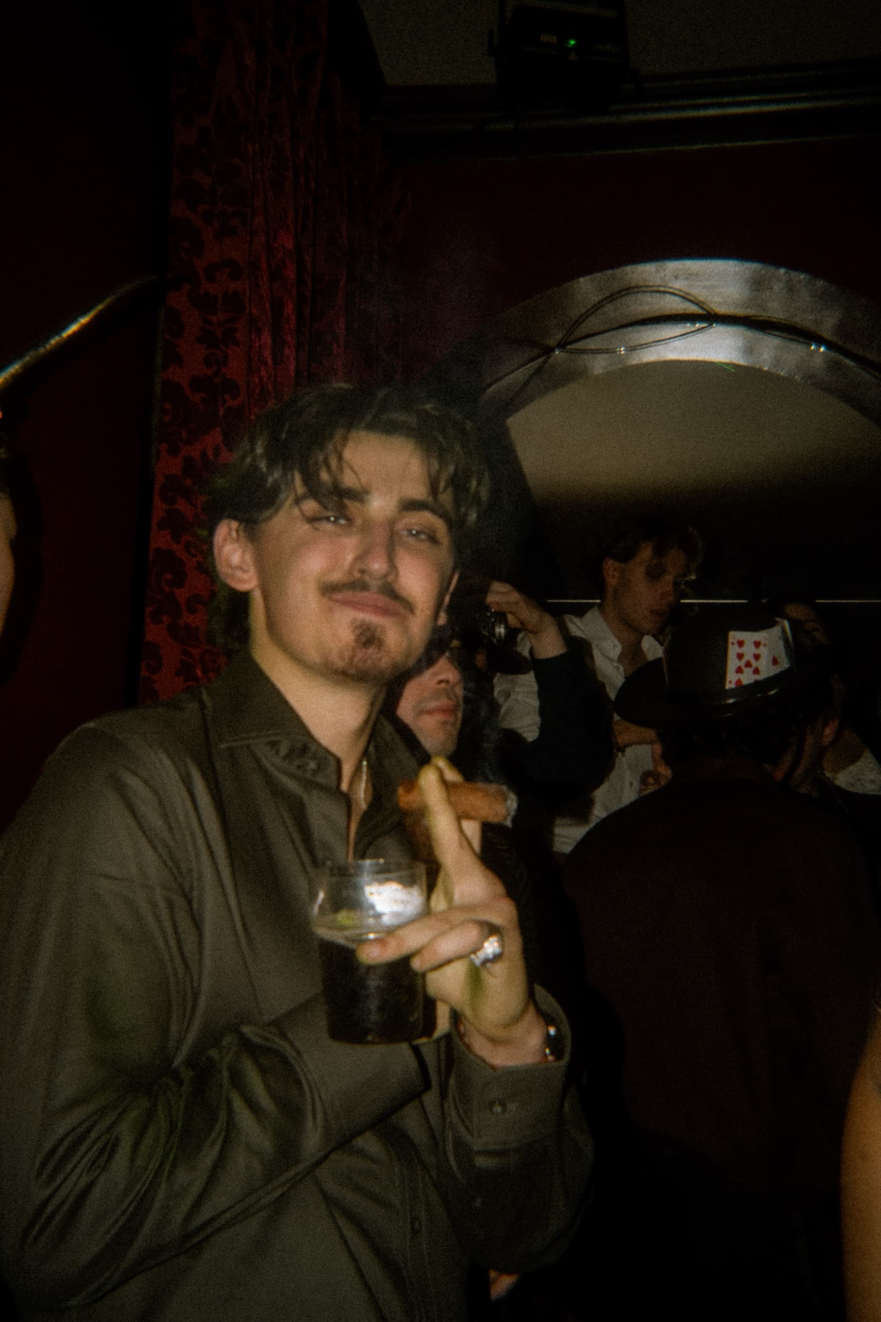 Guest holding a cocktail and smiling at camera against red damask wallpaper and velvet curtains