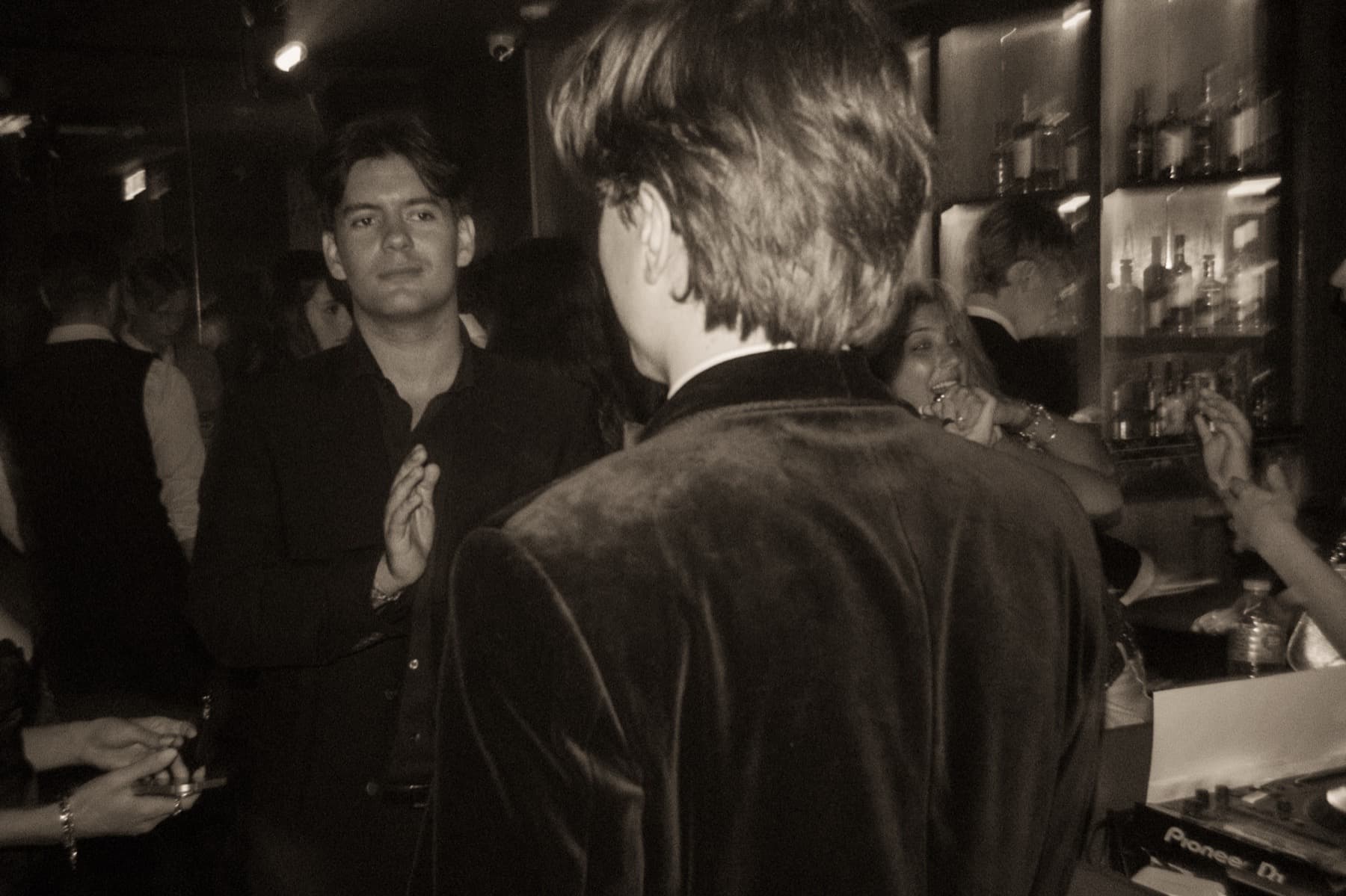 Two guests in velvet blazers chatting near the bar with bottles on illuminated shelves behind them