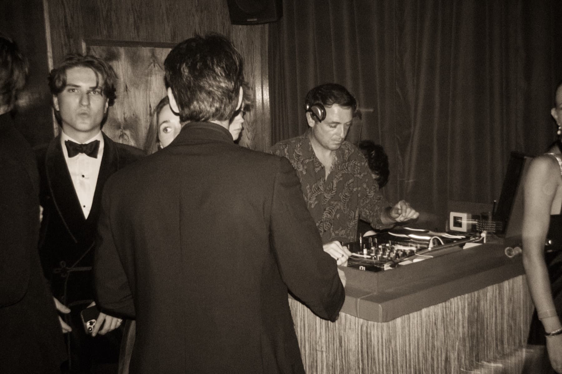 DJ in a patterned shirt with headphones adjusting the decks as guests in formalwear look on