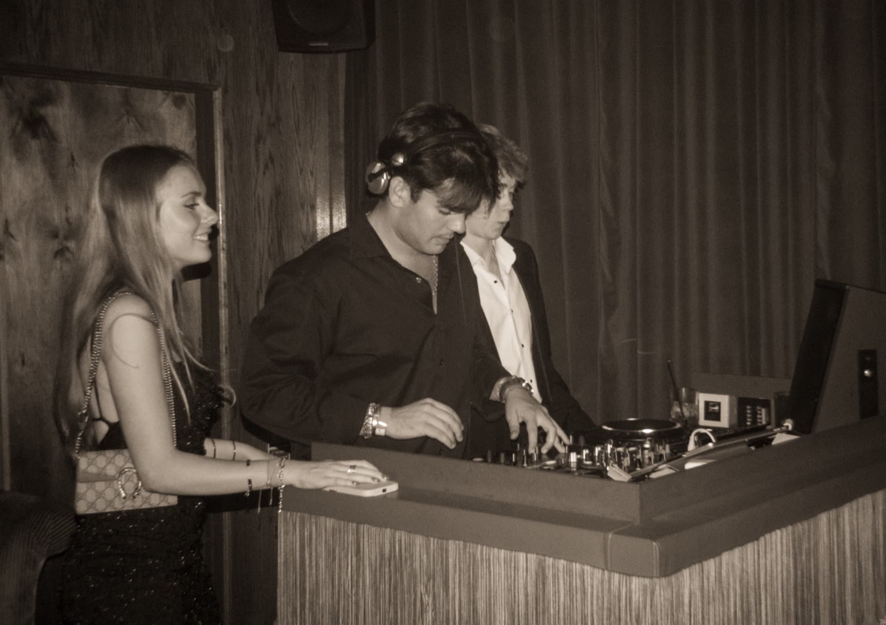 DJ mixing on Pioneer decks with headphones around his neck while a woman watches from beside the booth