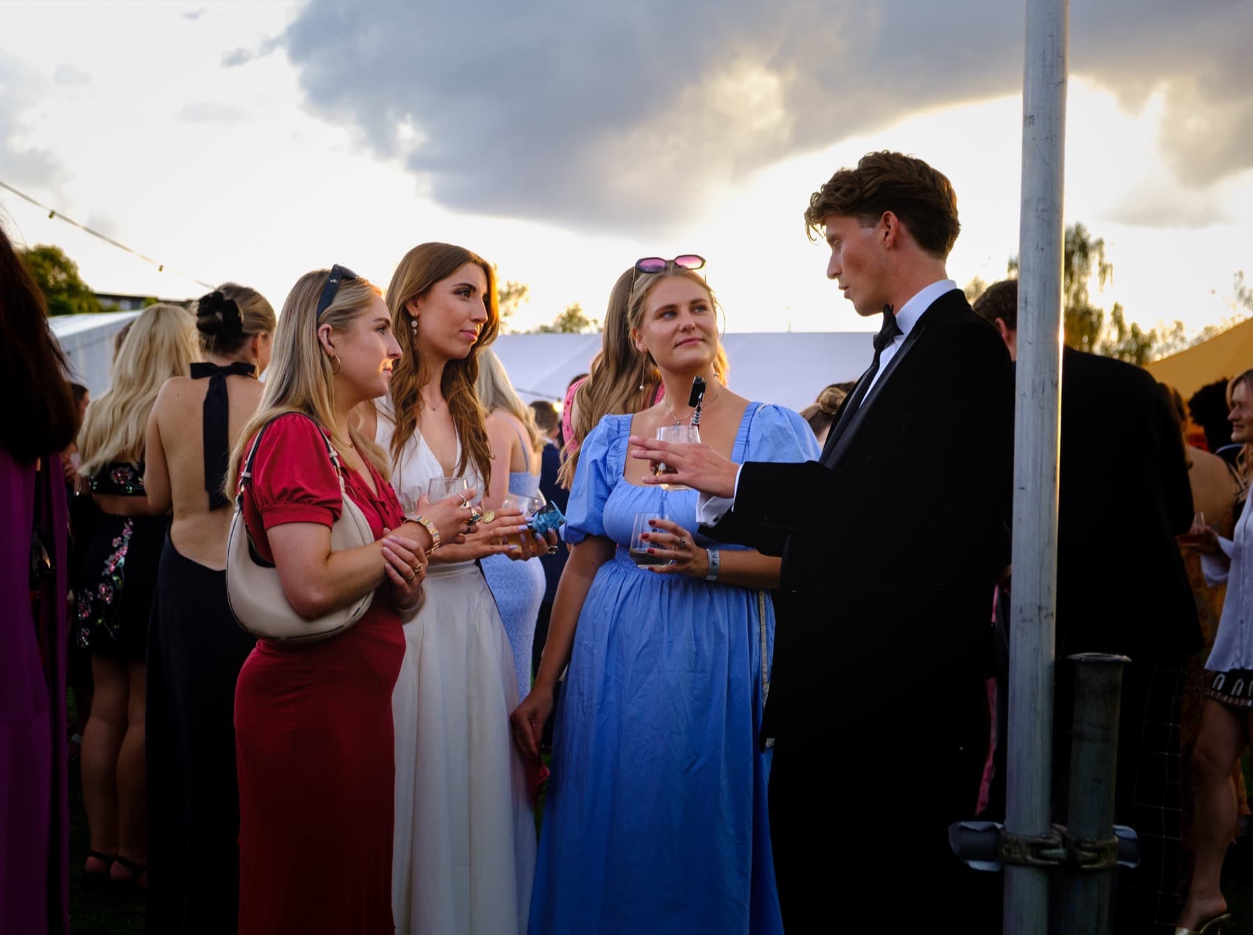 Group of friends in elegant dresses and black tie holding drinks and chatting at golden hour