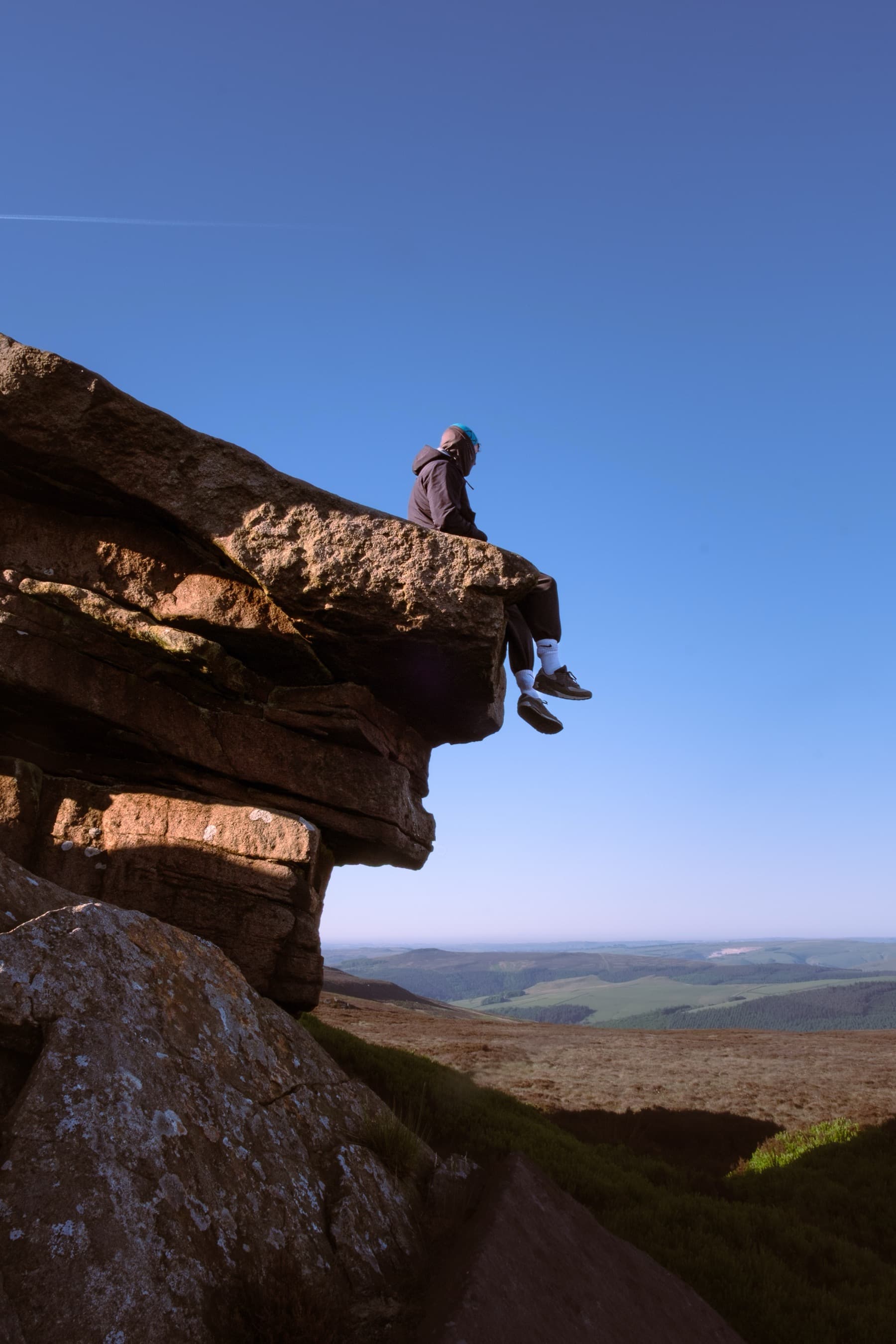 Person sitting on the edge of a gritstone tor with legs dangling over the drop, looking out over the moorland at golden hour