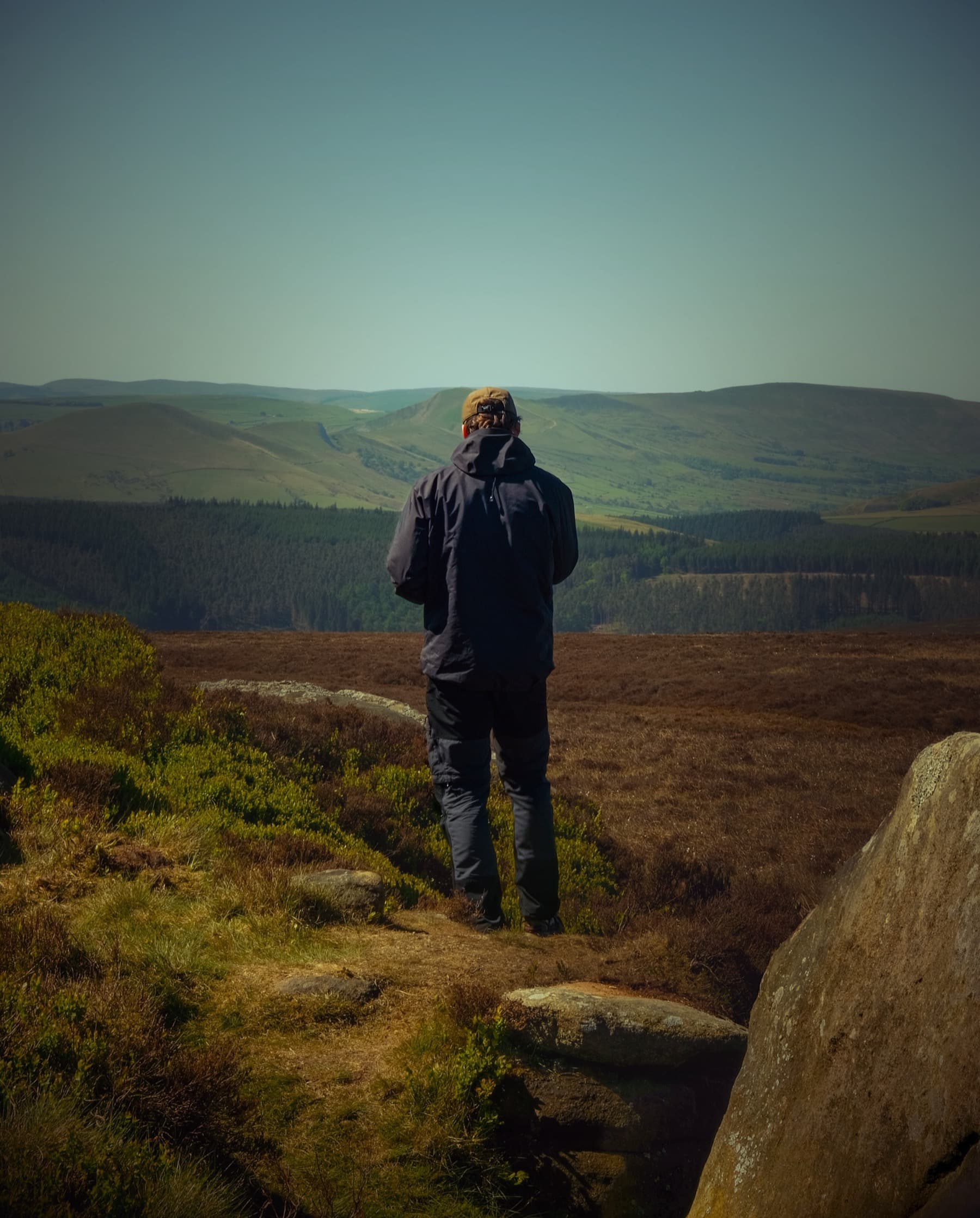 Hiker standing on a rocky outcrop looking across heather moorland and distant hills in warm afternoon light