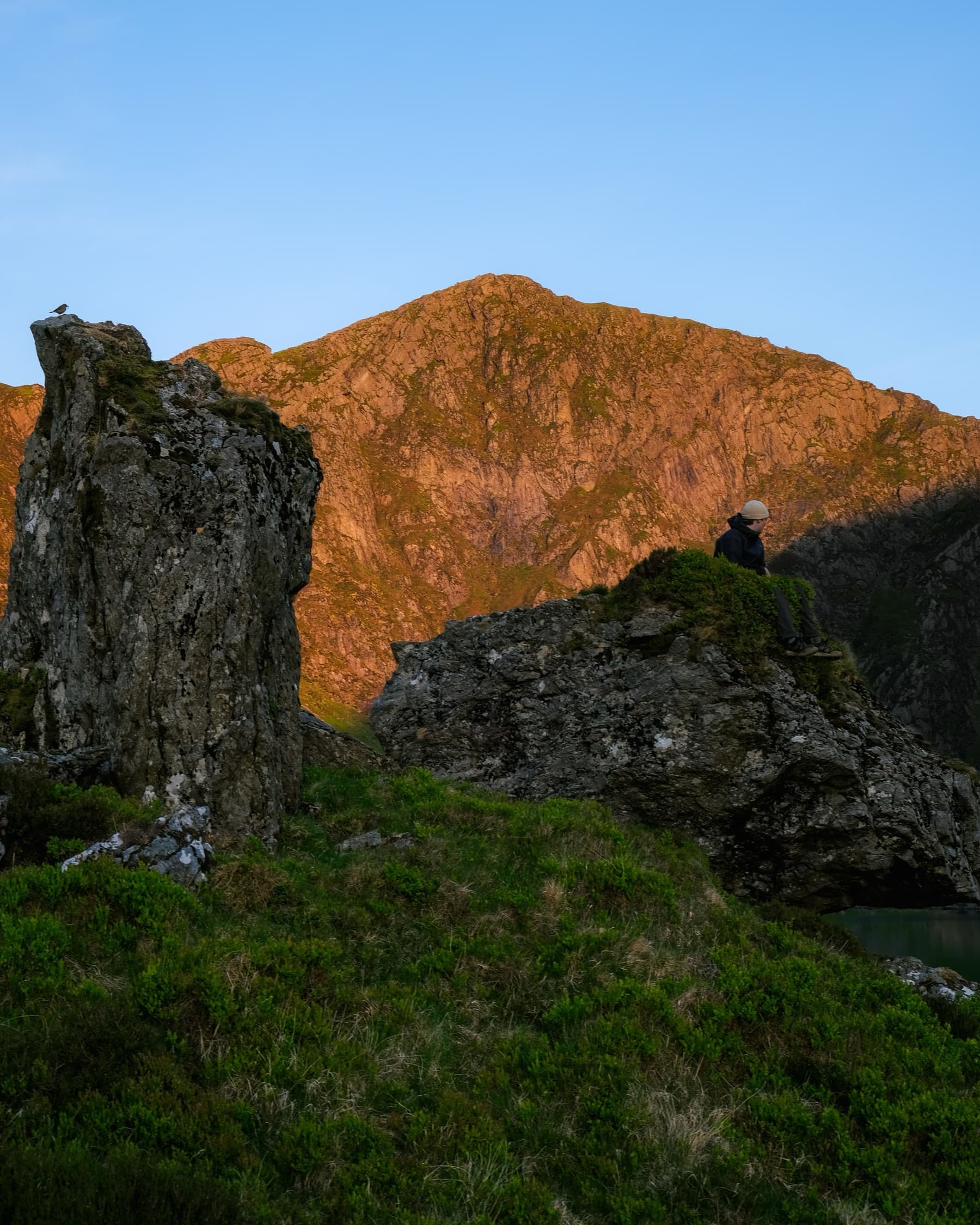 Person sitting among rock formations bathed in warm golden light with a sunlit mountain face behind at sunset