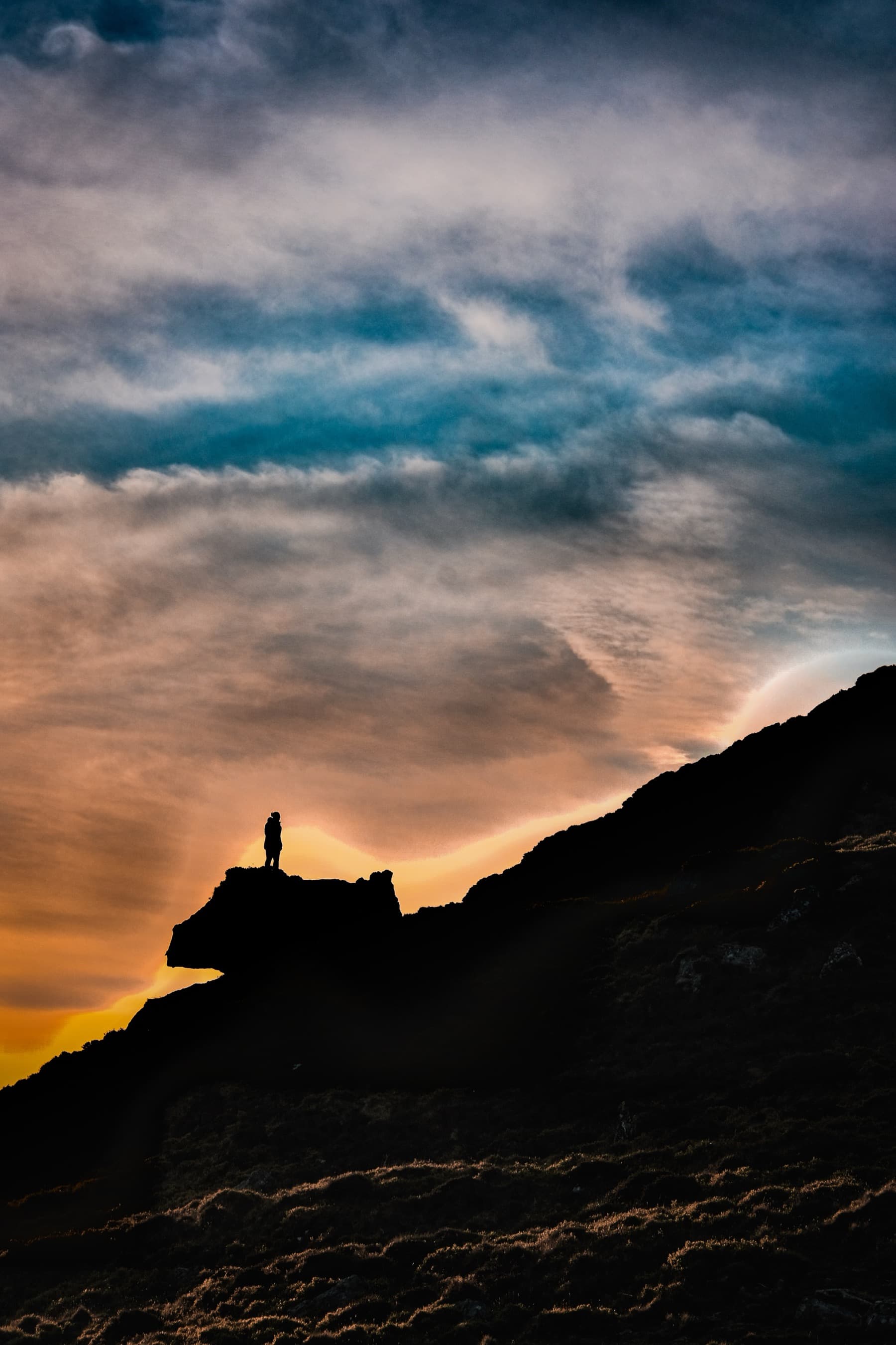 Silhouetted figure standing on a rock outcrop against a vivid sunset sky of orange, pink, and blue
