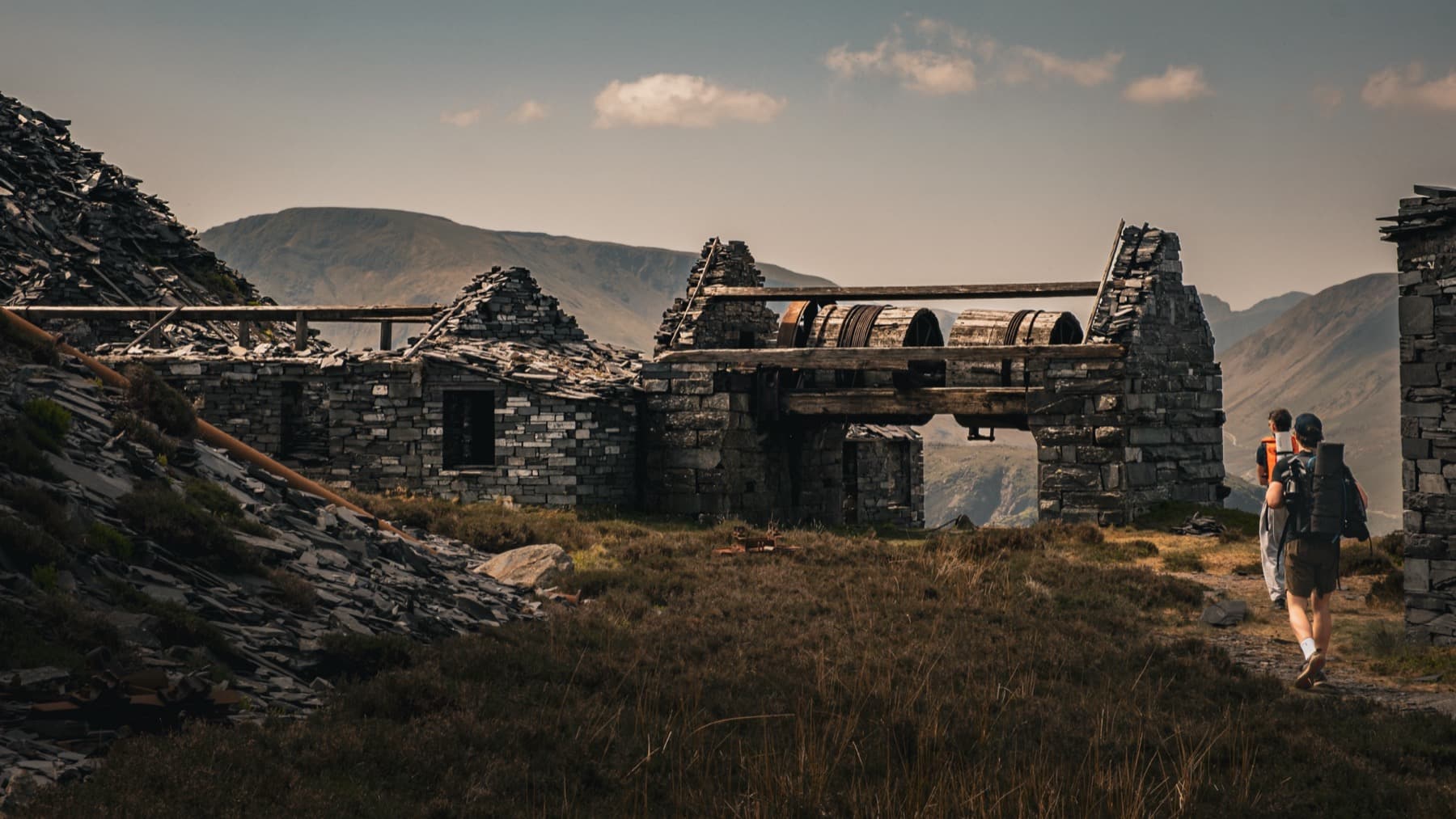 Hiker walking toward ruined quarry winding house with large iron drums and slate buildings in warm evening light