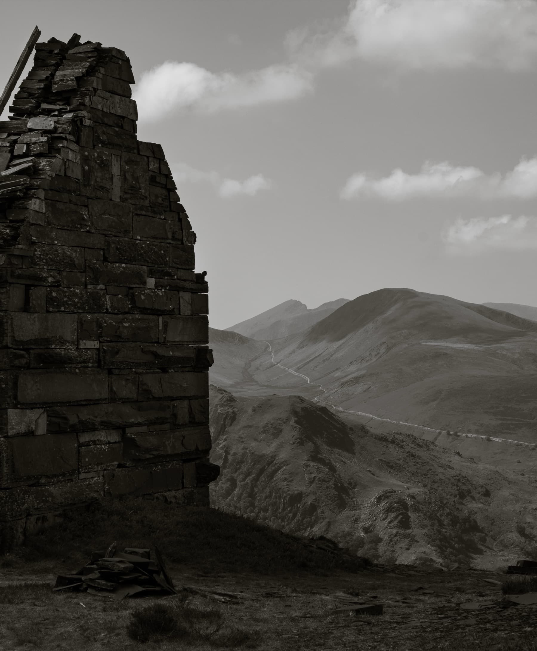 Ruined slate building gable end framing a distant mountain ridge and winding path in muted sepia tones