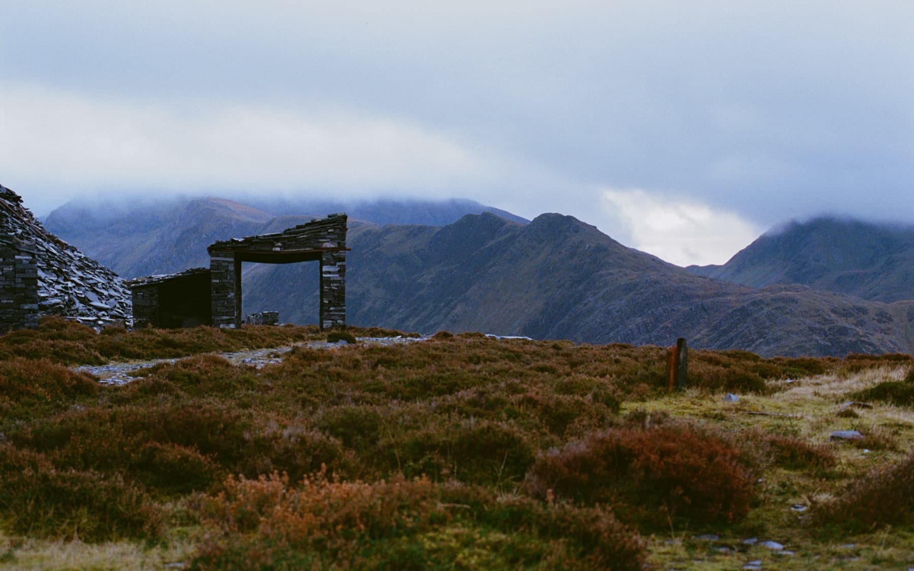 Ruined quarry shelter with open doorway framing cloud-wrapped Snowdonia peaks across a heather moorland, shot on film