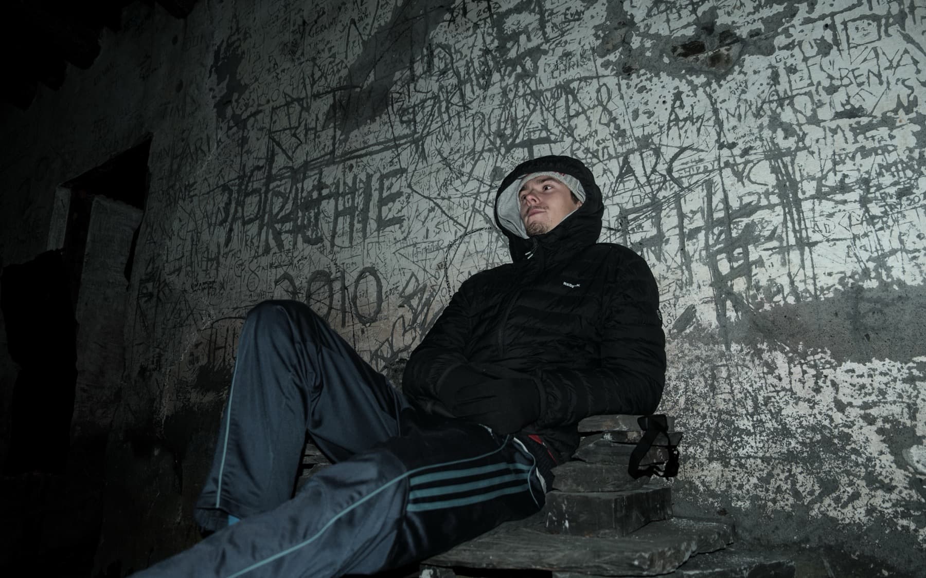 Person resting against graffiti-covered quarry shelter walls lit by flash in the darkness