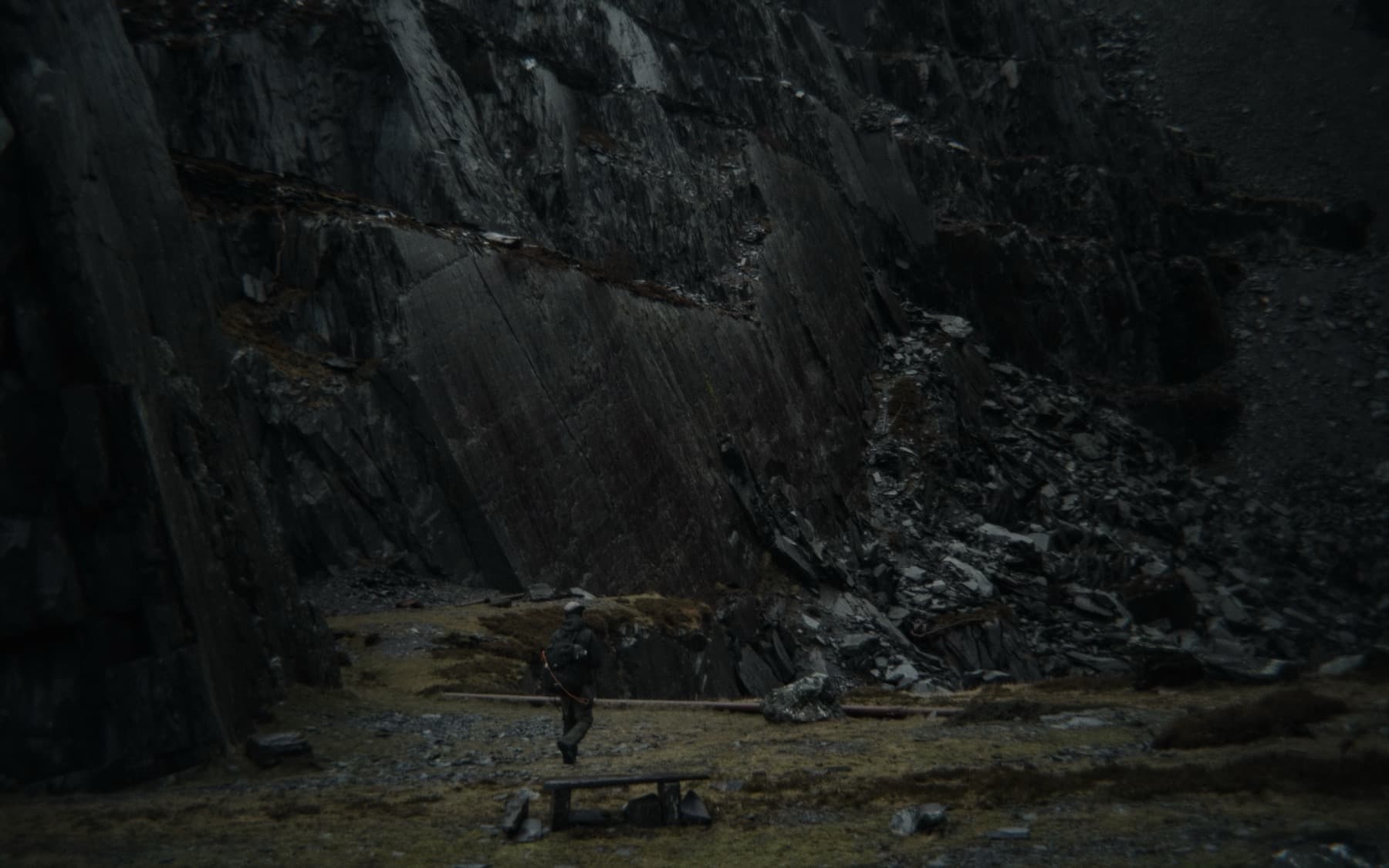 Lone hiker dwarfed by a towering dark slate quarry face with a bench in the grassy foreground