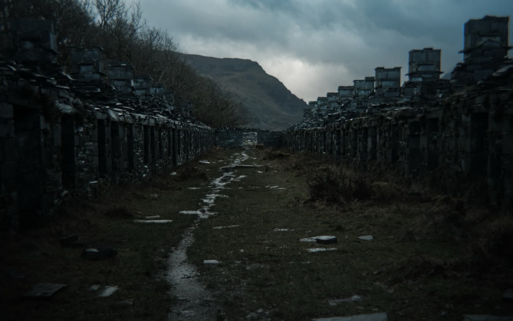 Rows of ruined slate quarry workers' barracks lining a muddy path with a mountain rising beyond under stormy skies