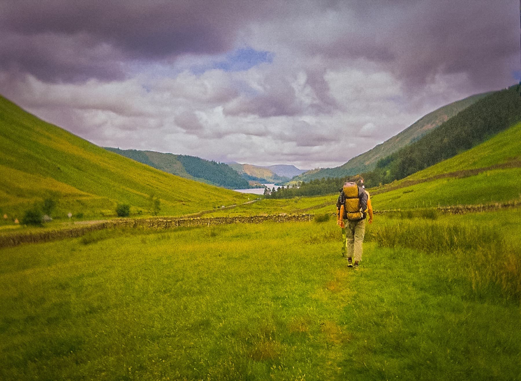 Hiker with a large backpack walking through a lush green valley toward distant hills and a lake, shot on film
