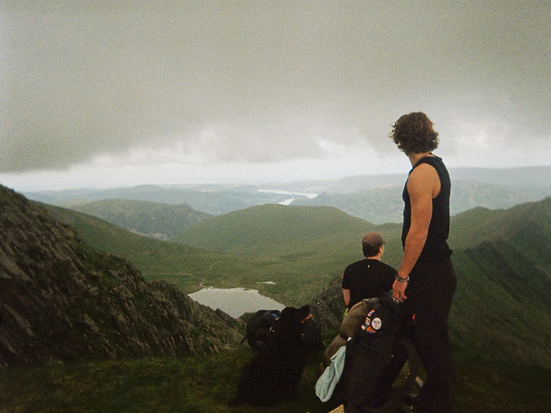 Two hikers standing on a summit looking out over green valleys and lakes under an overcast sky, shot on film
