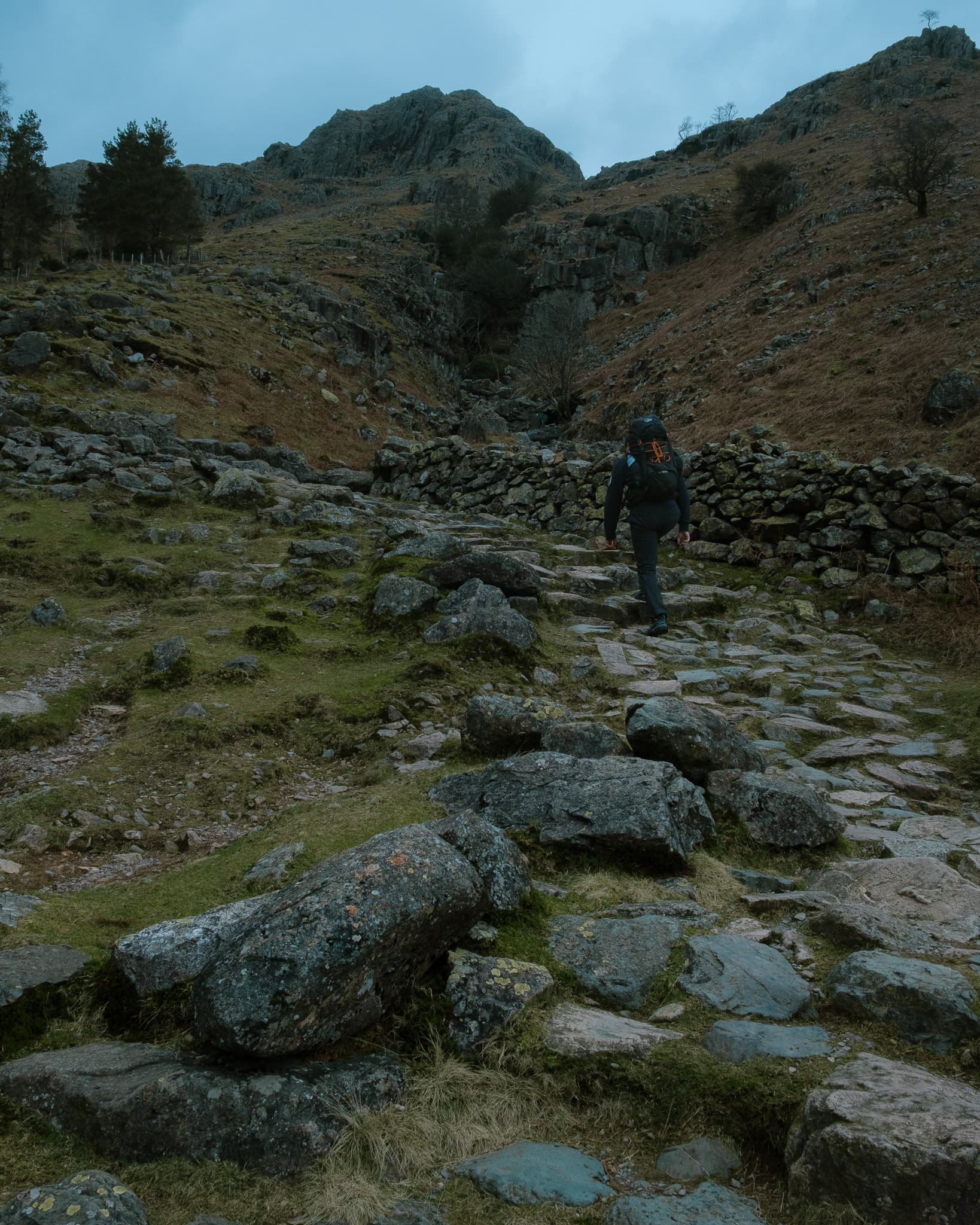 Hiker walking up a rocky stone path toward craggy peaks with a dry stone wall and moody overcast sky