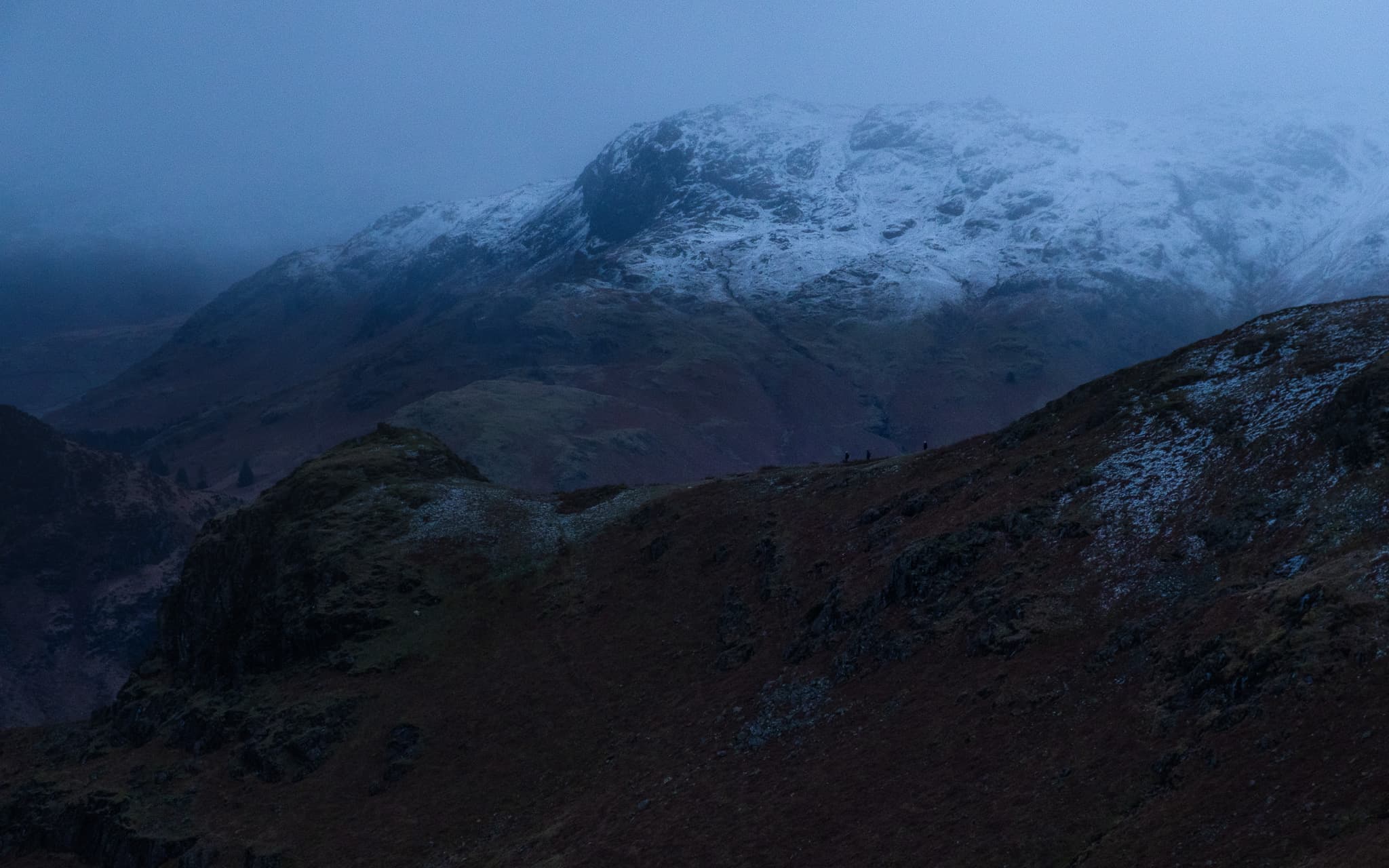 Snow-covered Langdale Pikes rising into low cloud and mist with dark moorland ridges in the foreground