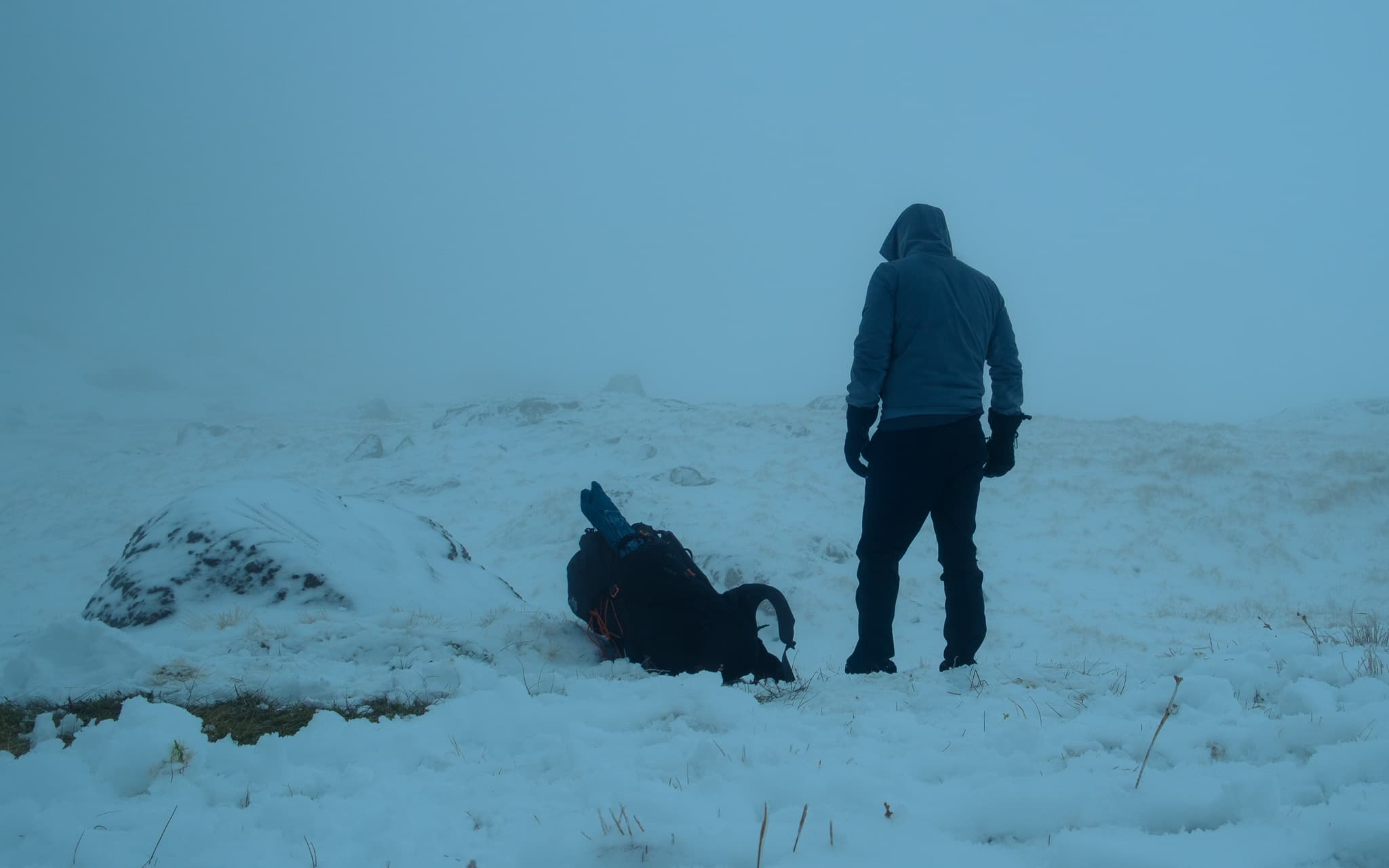 Hiker standing on a snowy summit looking into thick fog with a backpack at their feet