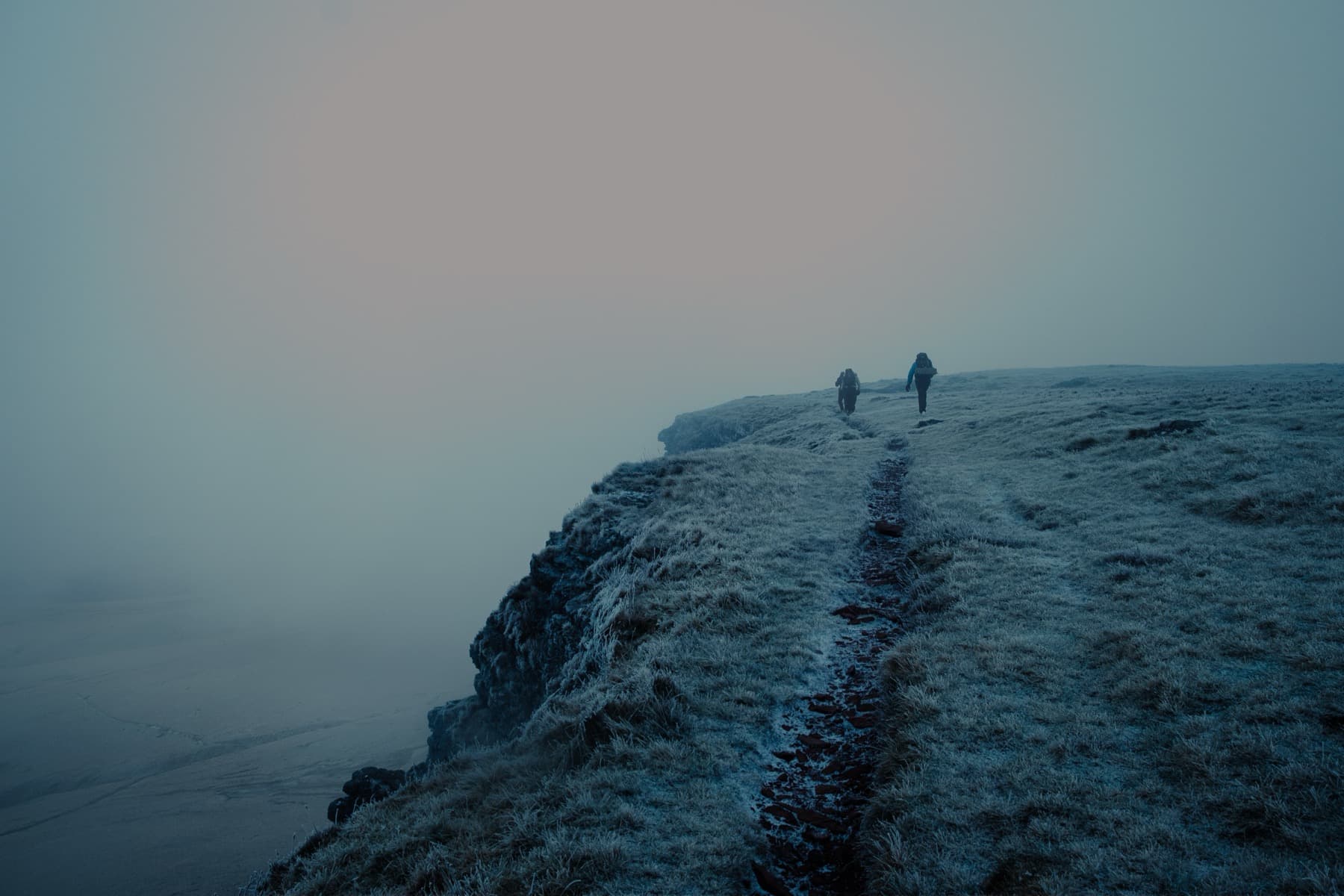 Two hikers walking along a frosty cliff-edge path above a steep drop into thick mist