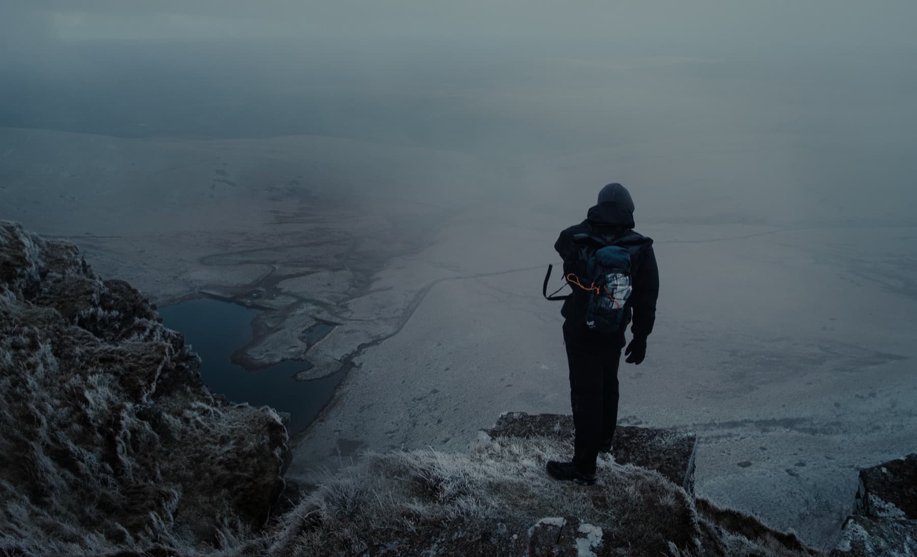 Hiker standing on a frosty ridge edge looking down over a frozen landscape and distant lake in heavy mist