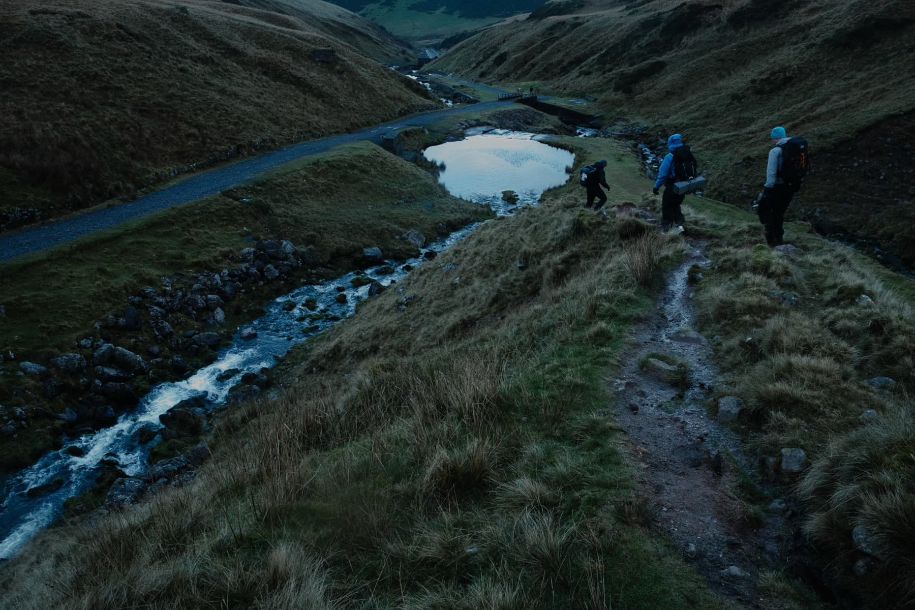 Three hikers descending a hillside trail beside a stream toward a small pool with mountains behind at dusk