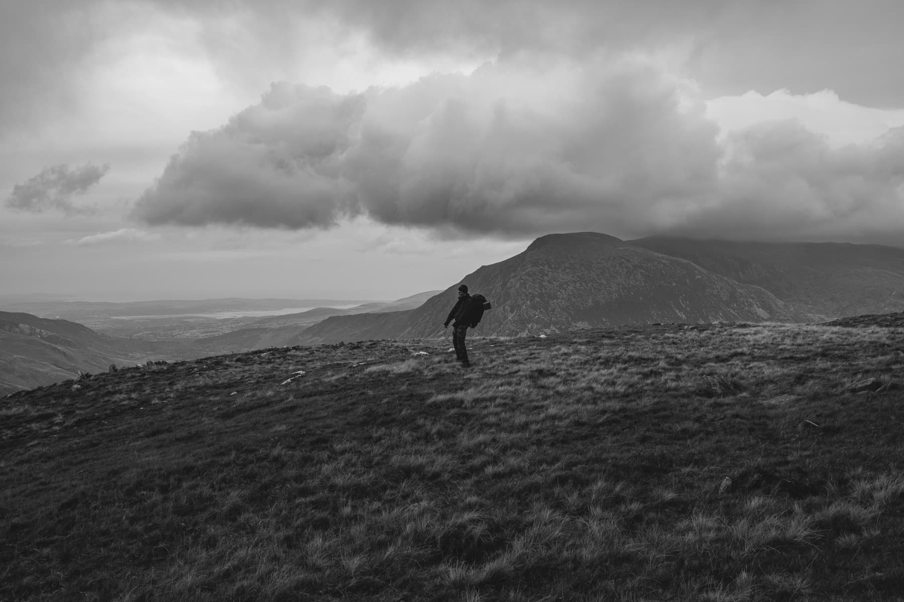 Lone hiker walking across windswept moorland in black and white with a mountain backdrop and billowing clouds