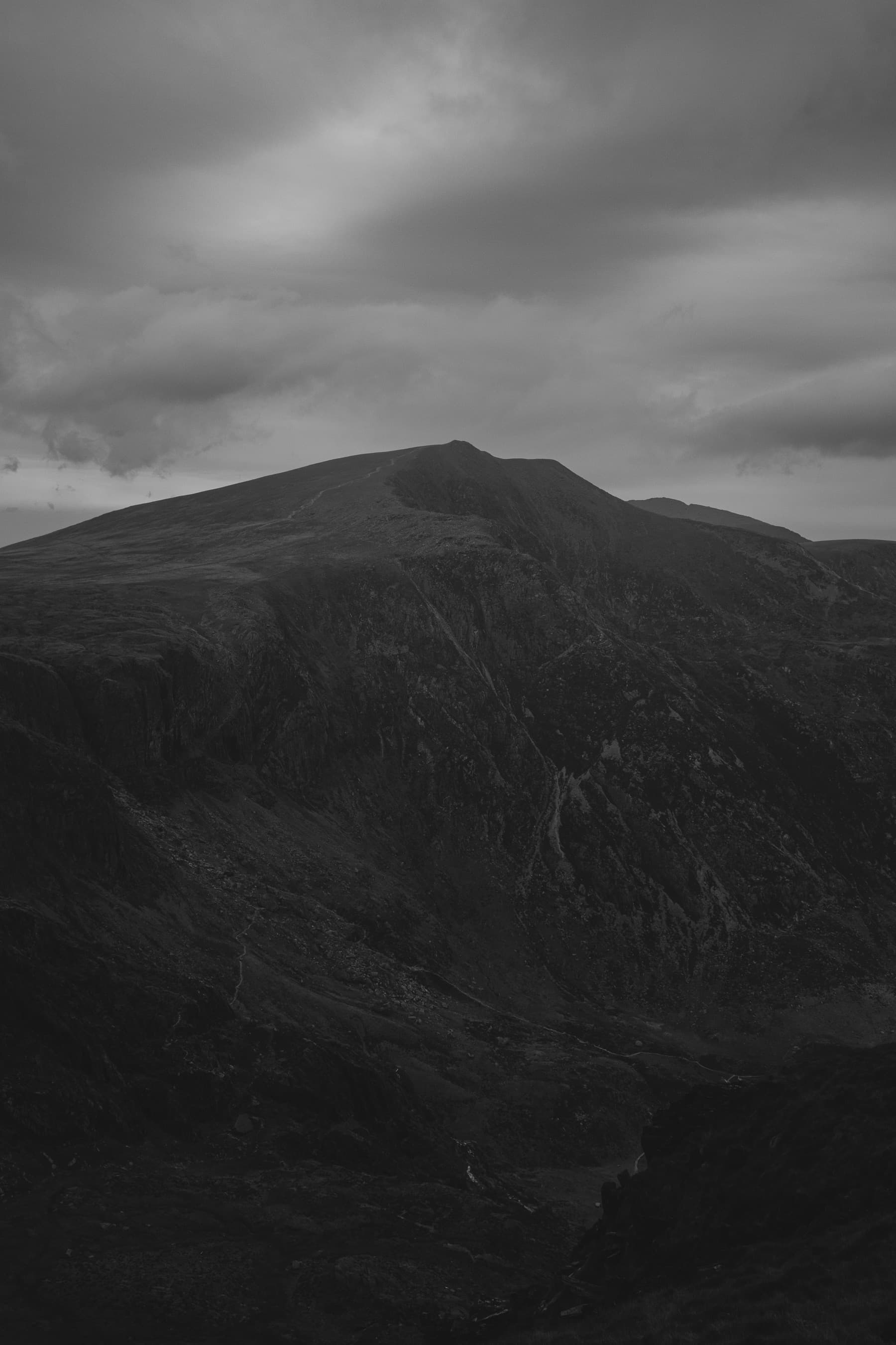 Mountain summit in black and white rising above a dark cwm with a path visible along the ridge under moody clouds