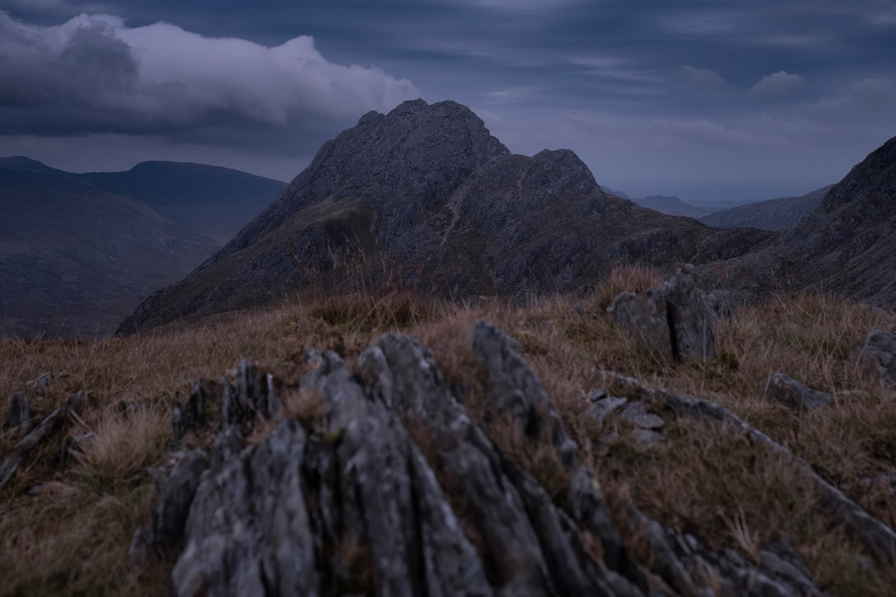 Jagged rocky peak of Tryfan rising dramatically against dark clouds with golden grass and slate in the foreground