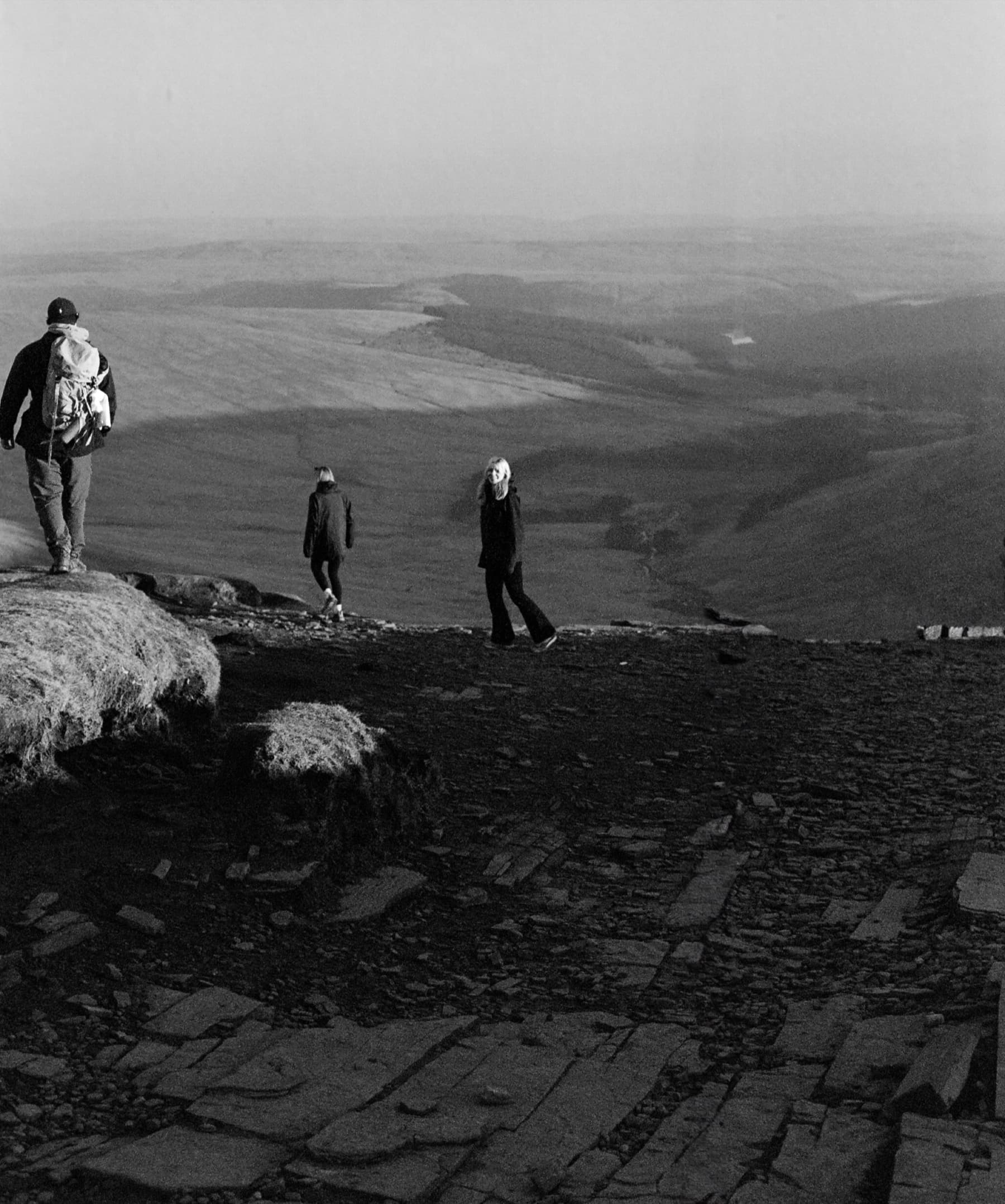 Three hikers descending the rocky summit plateau of Pen y Fan with sweeping valley views behind in black and white film