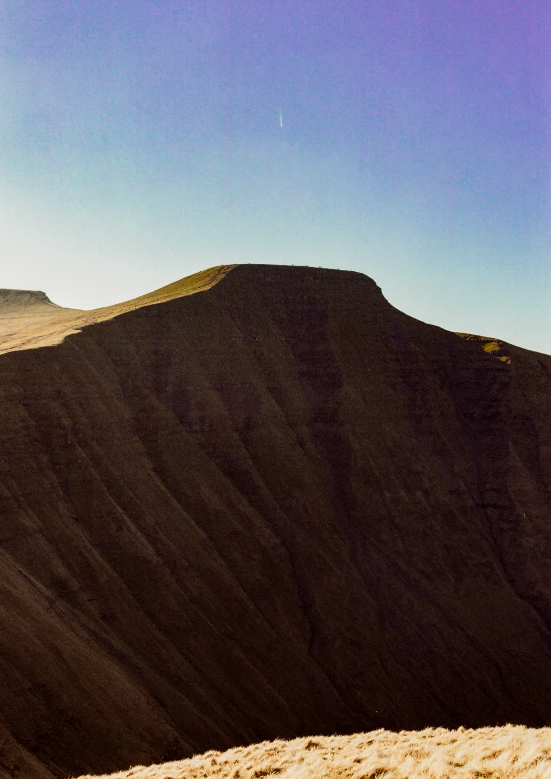 The steep north face of Pen y Fan rising sharply against a clear blue sky with dry grass in the foreground, shot on film