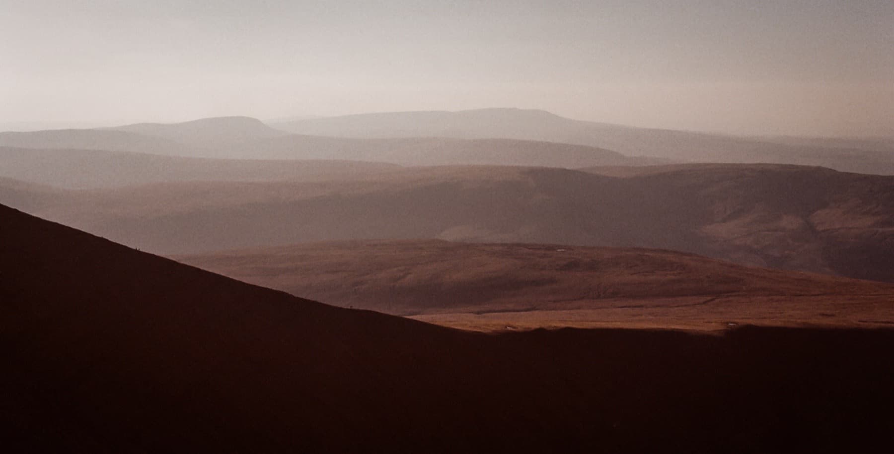 Layered mountain ridges receding into warm hazy light at golden hour with tiny figures silhouetted on the nearest ridge, shot on film