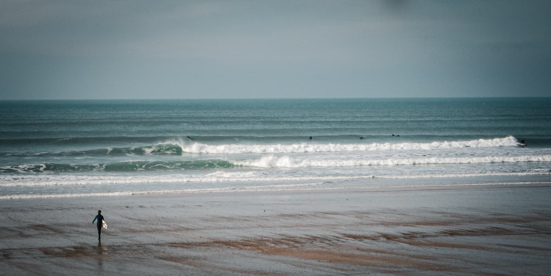 Lone surfer carrying a board walks across a vast wet beach toward lines of breaking waves with other surfers in the water