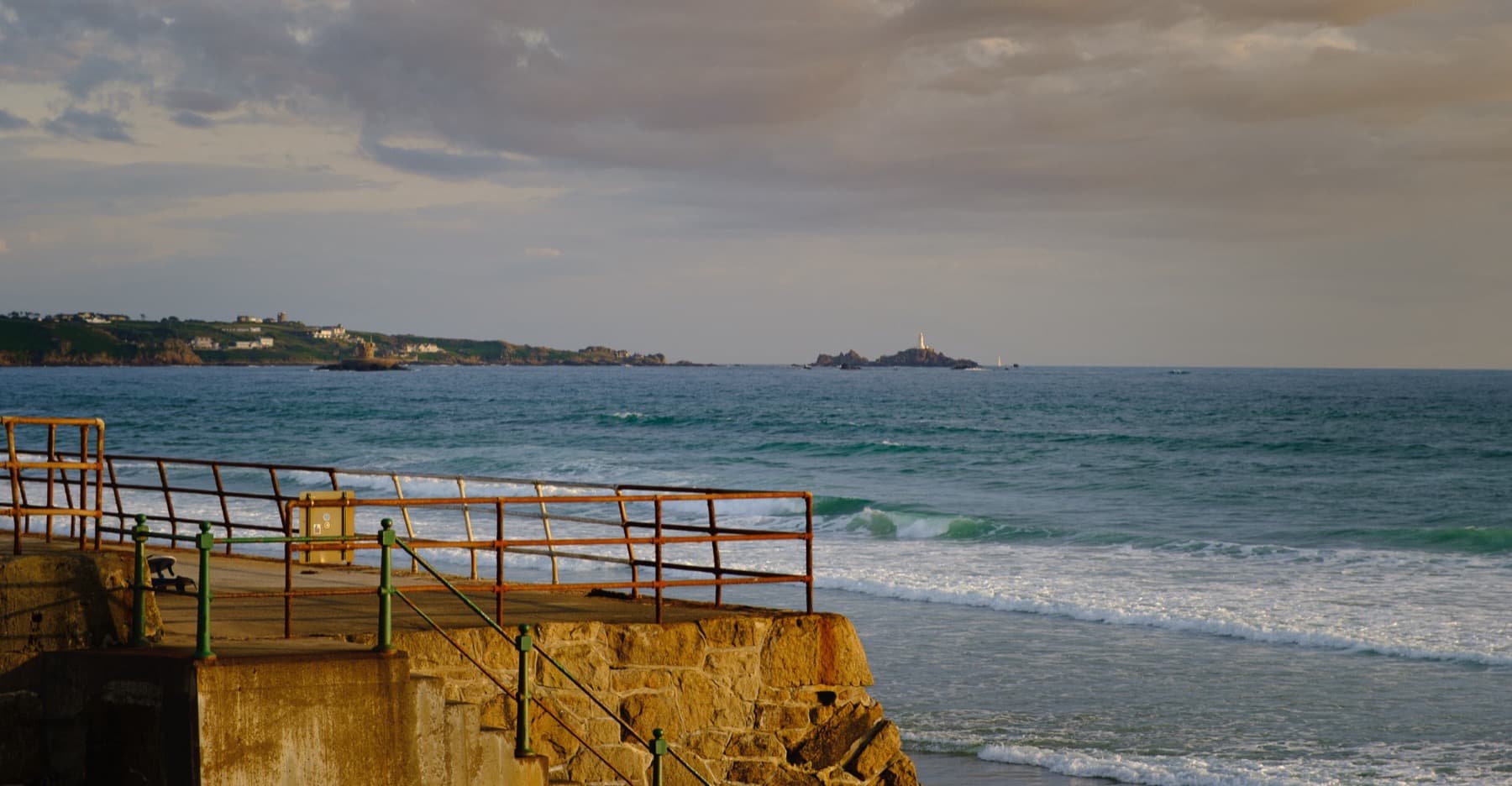 Stone sea wall with rusted metal railings overlooking turquoise waves, with a distant lighthouse and headland under golden evening clouds