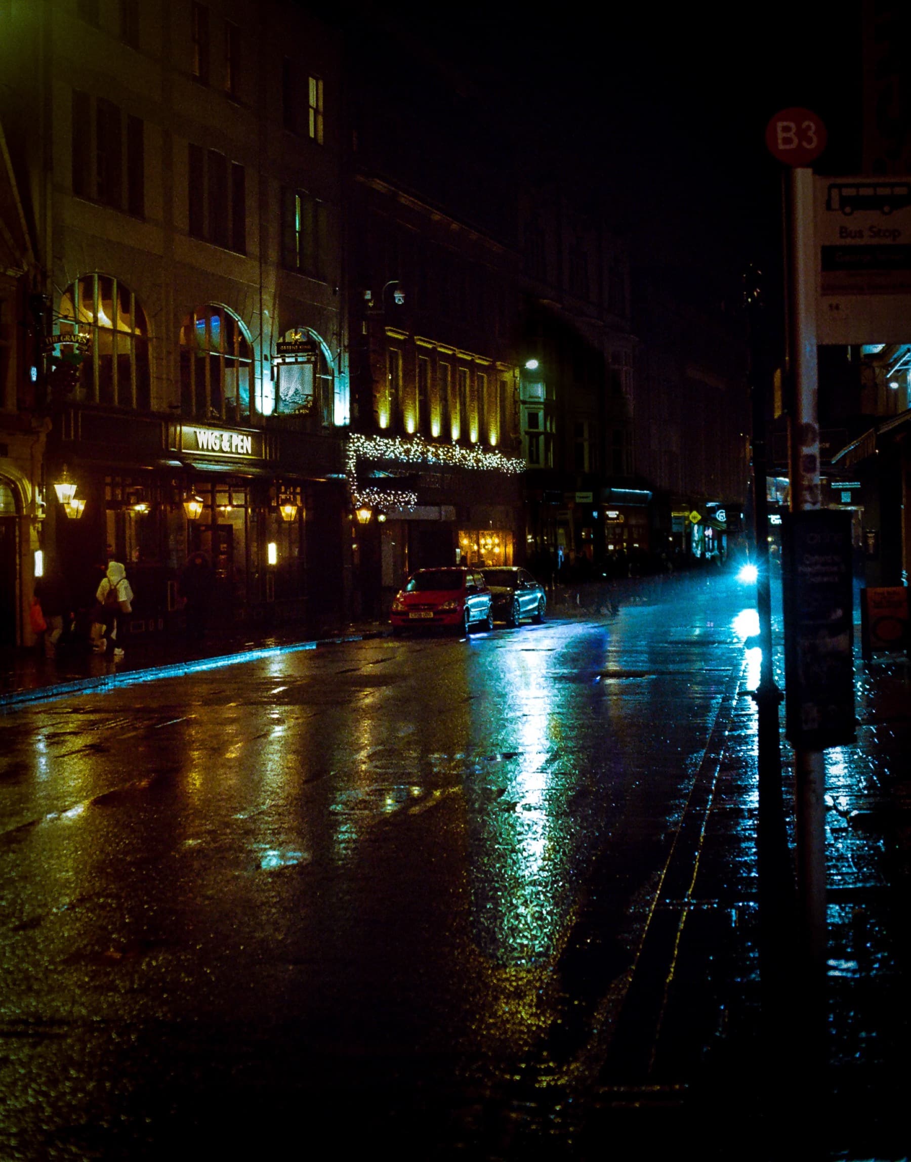 Rain-slicked London street at night with warm pub lights and coloured reflections on wet pavement