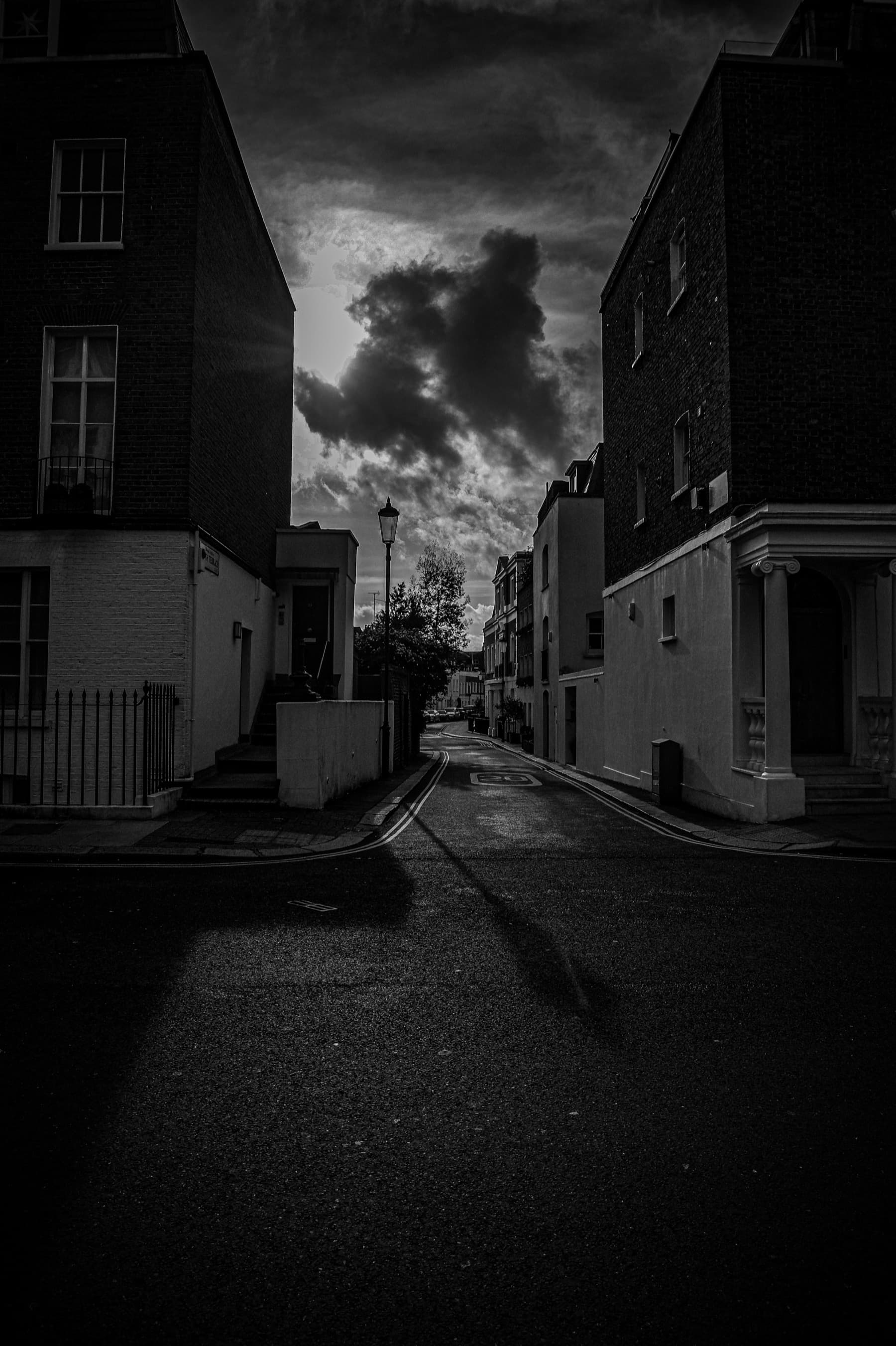 Dramatic black and white view down a narrow London mews with dark shadows framing a moody sky and distant street lamp