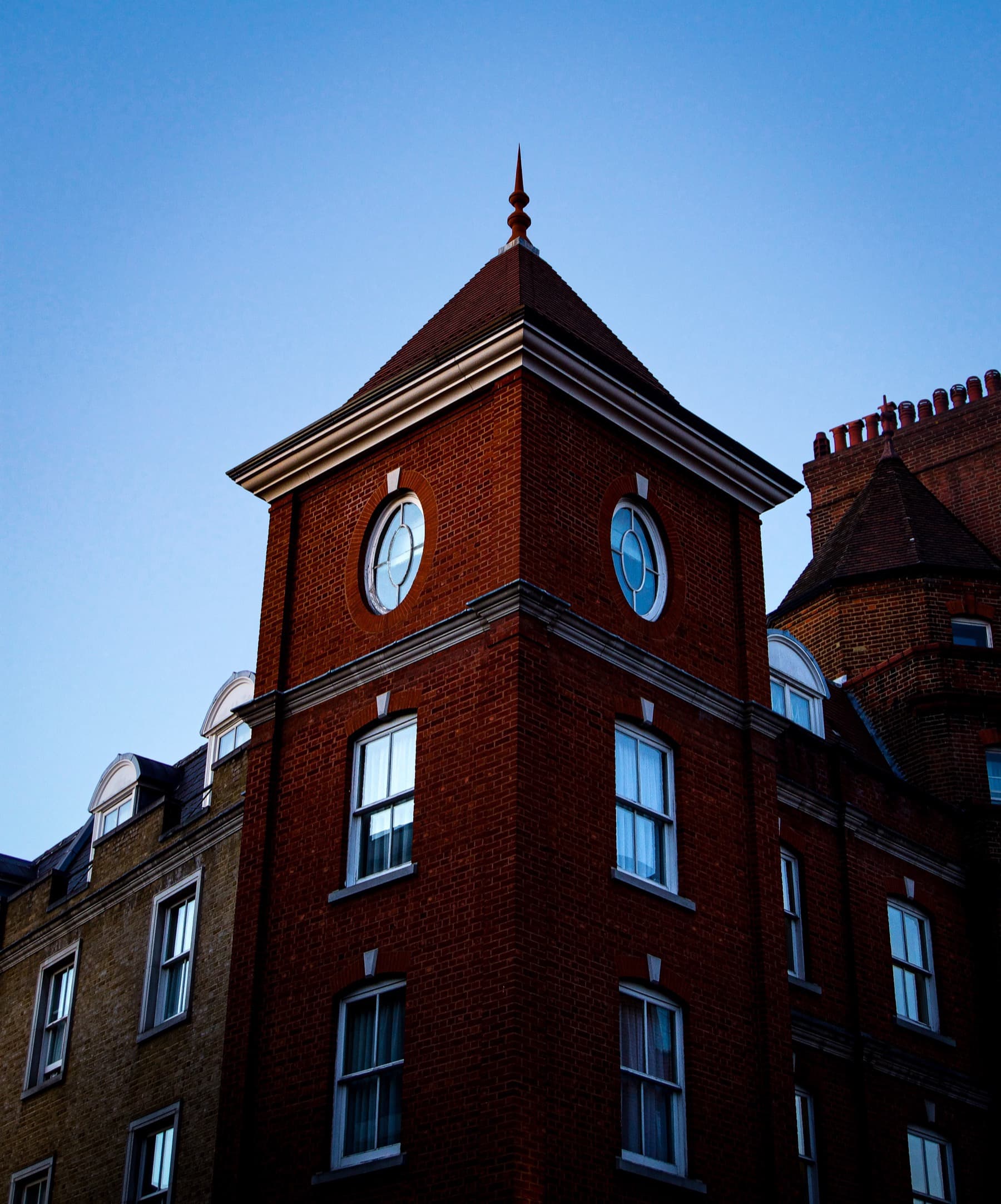 Red brick Victorian tower with round windows and pointed spire against a clear blue dusk sky