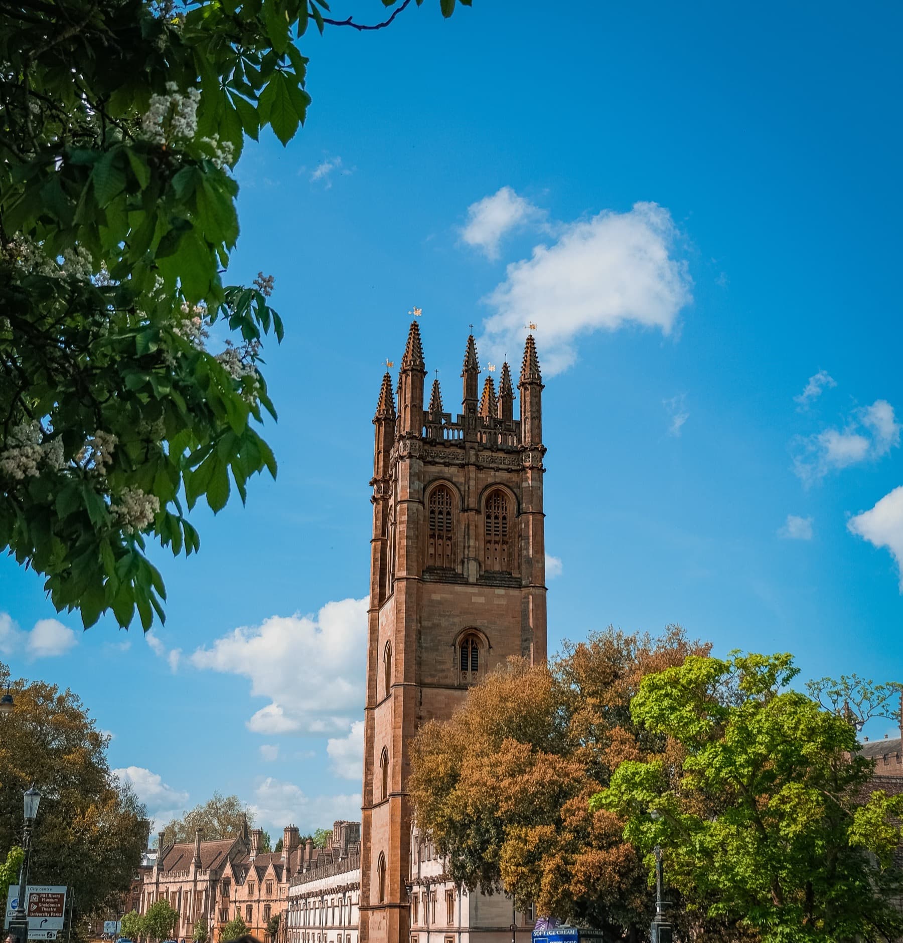 Magdalen College tower in Oxford framed by green trees against a bright blue sky with white clouds