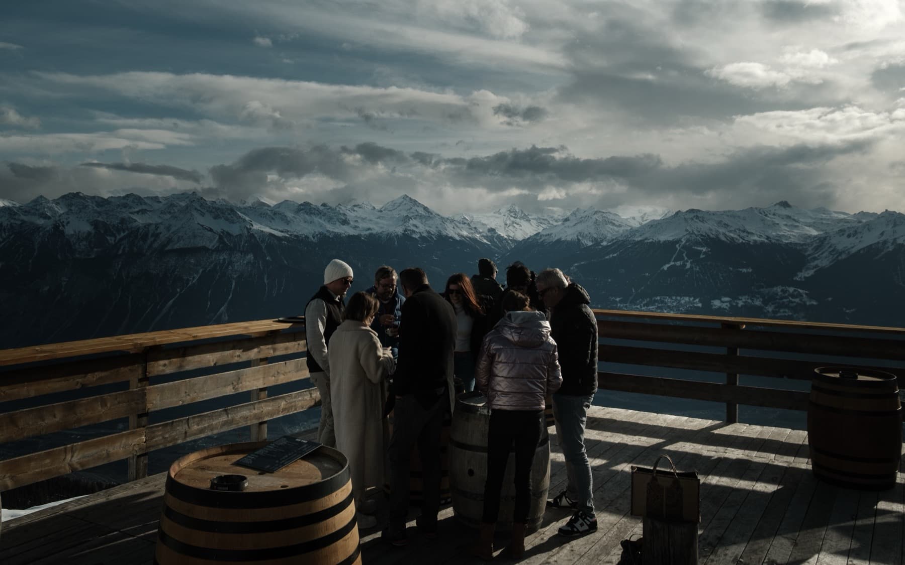 Group of people gathered around wine barrels on a wooden mountain terrace, dramatic snow-capped peaks and stormy clouds filling the background