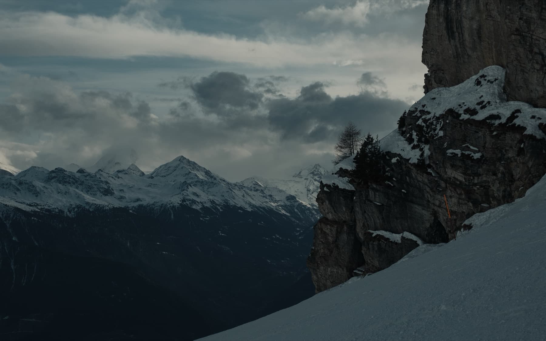 Steep rocky cliff face with a lone tree clinging to the edge, snow-covered peaks visible through clouds in the distance under a dark moody sky