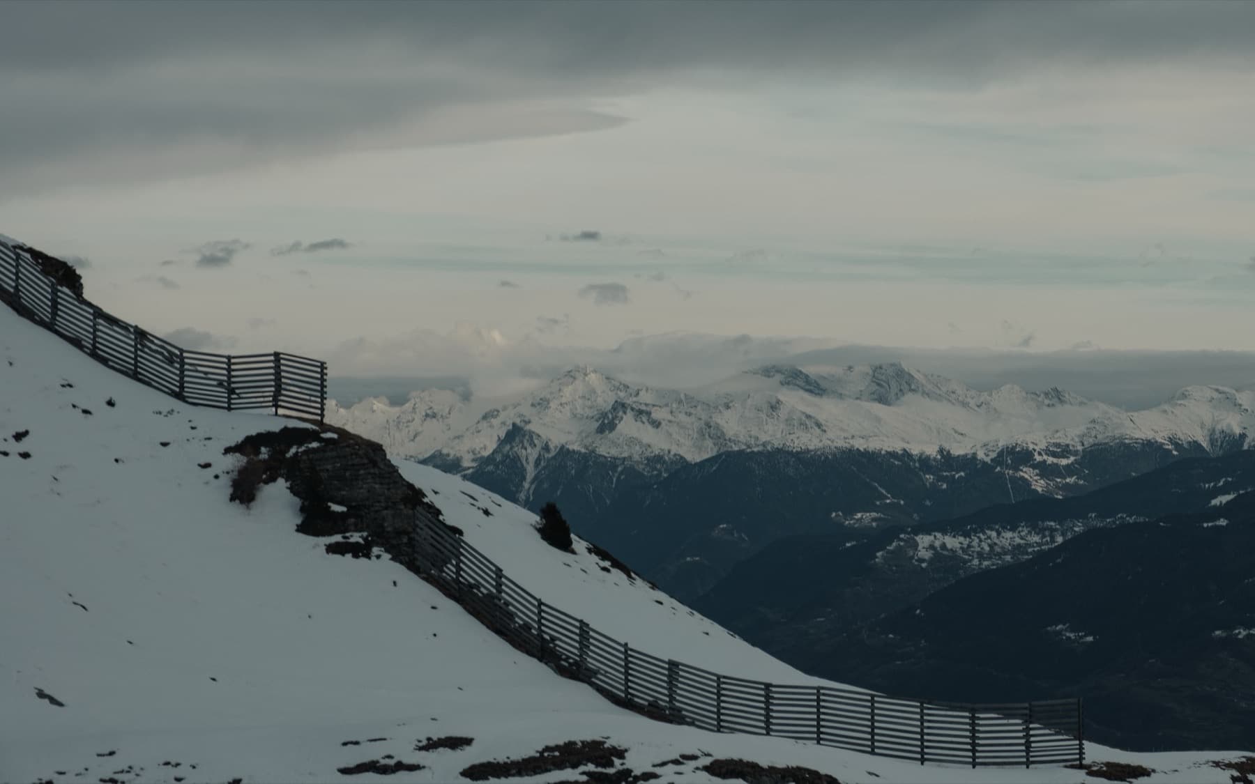 Avalanche barrier fence curving along a snowy ridgeline with a vast panorama of alpine peaks fading into cloud and haze beyond