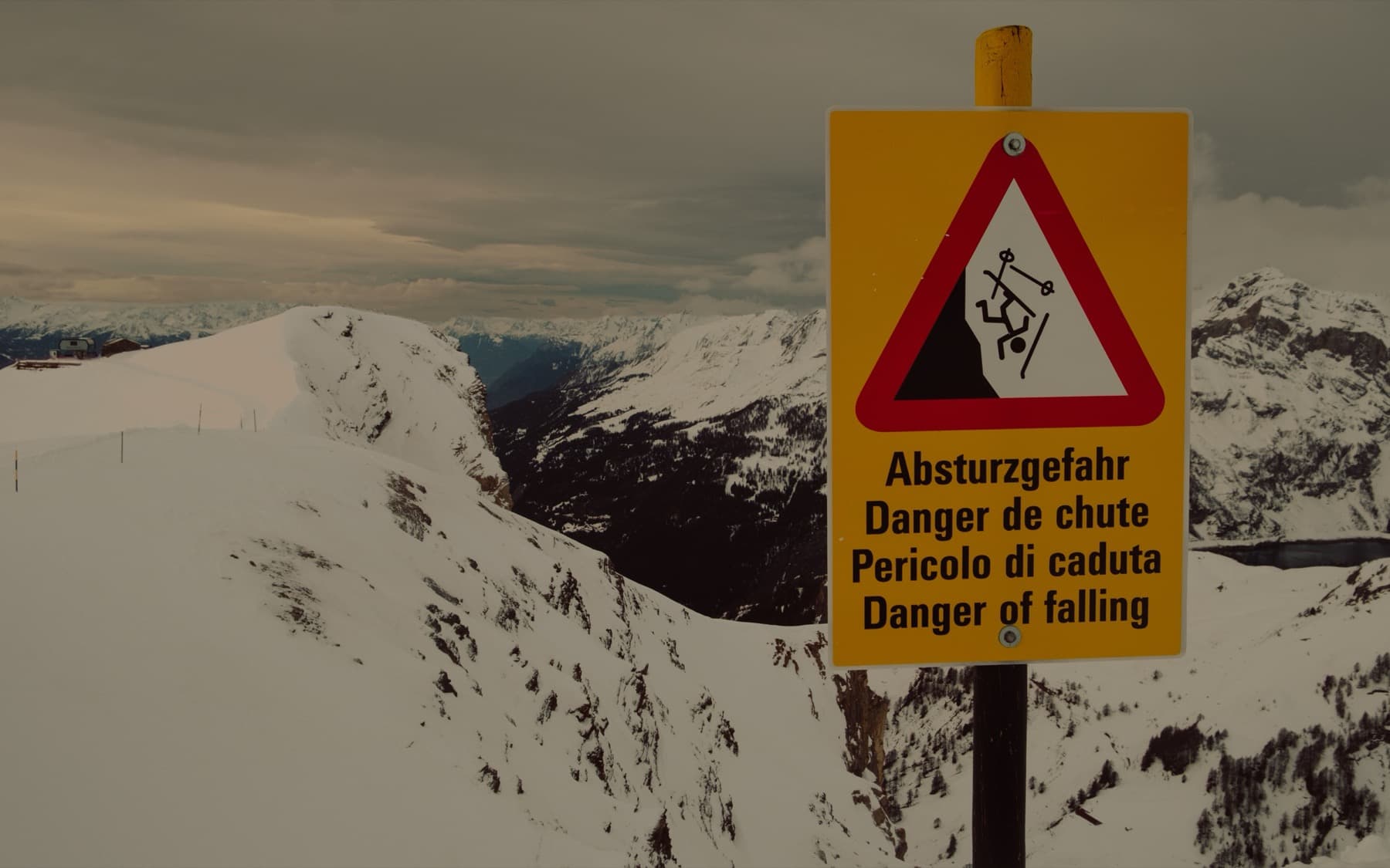 Multilingual danger of falling warning sign on a snowy mountain ridge with steep drop-offs and alpine peaks under overcast skies behind