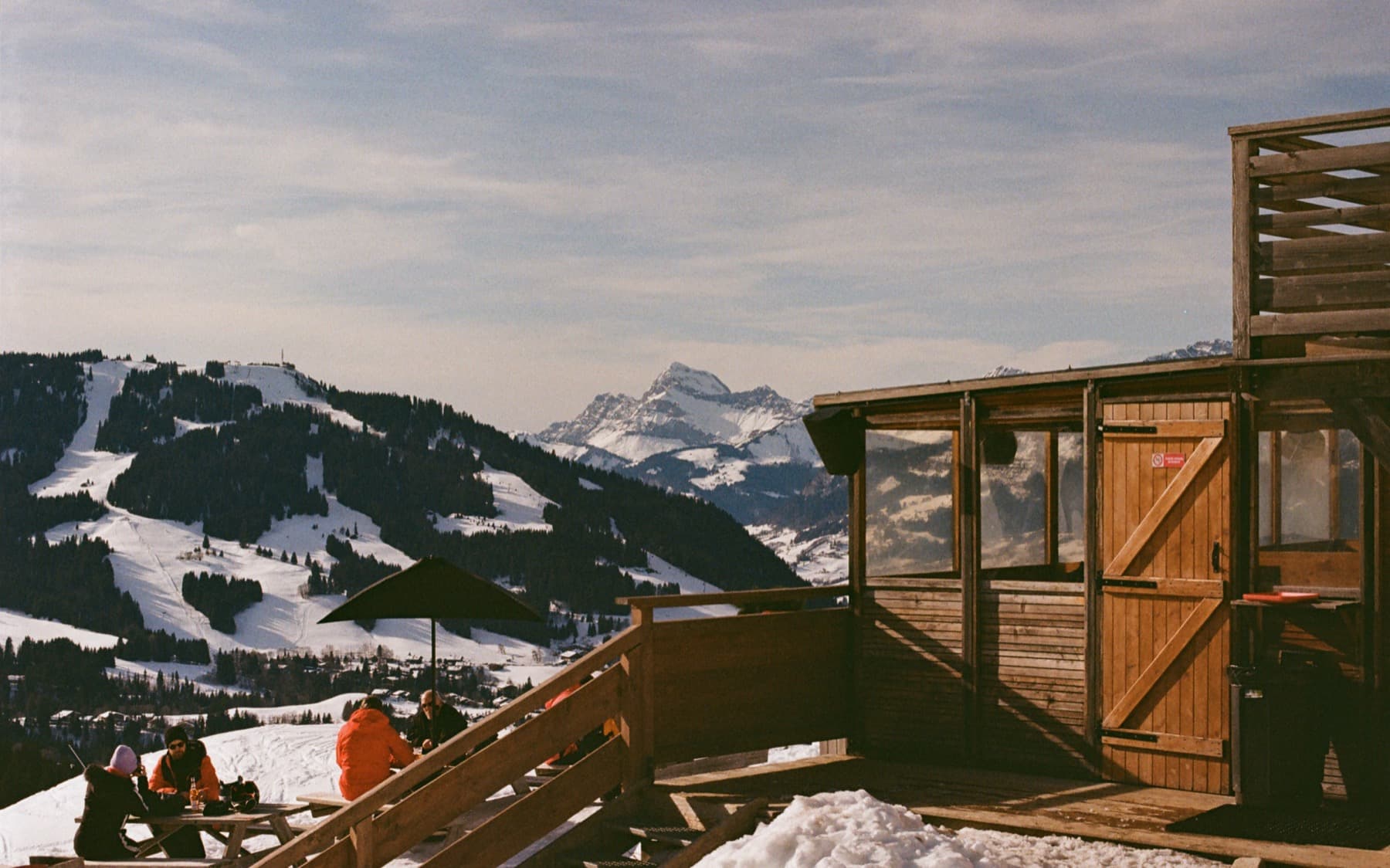 Wooden mountain hut terrace with skiers resting beside it, overlooking snow-covered slopes and a jagged peak in the distance