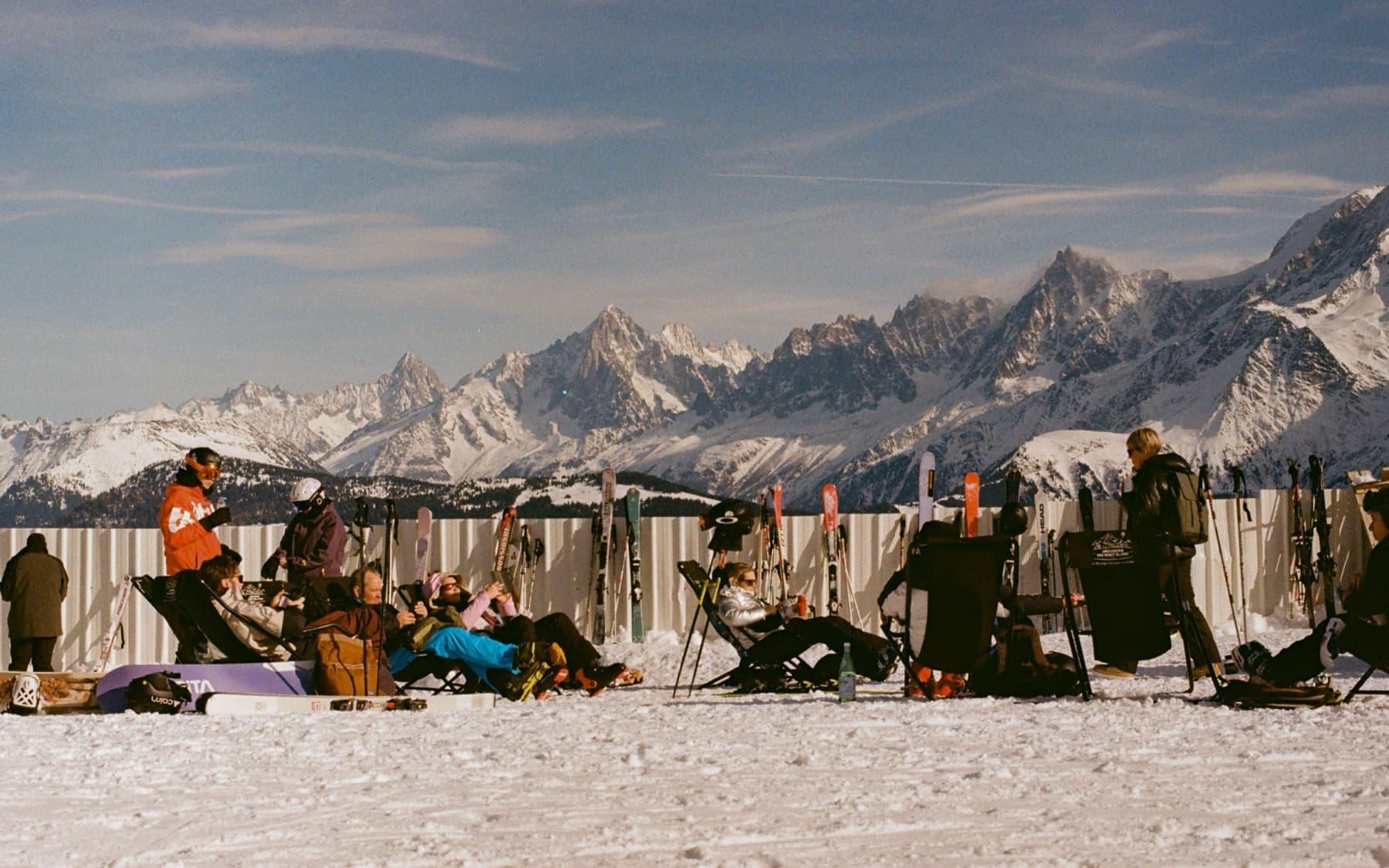 Skiers relaxing in deck chairs at a mountaintop terrace with skis and snowboards propped up, sharp alpine peaks stretching across the horizon