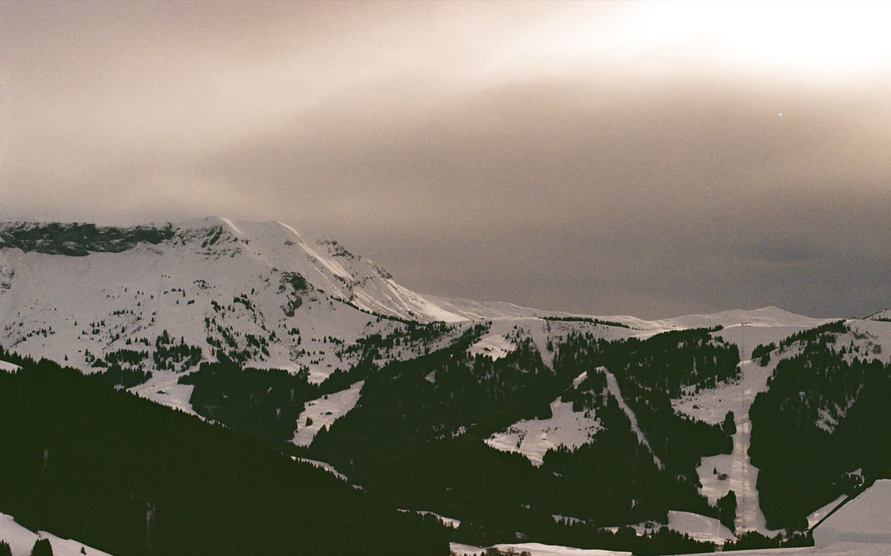 Moody overcast sky above snow-dusted mountain ridges with dark forested valleys and ski runs visible below
