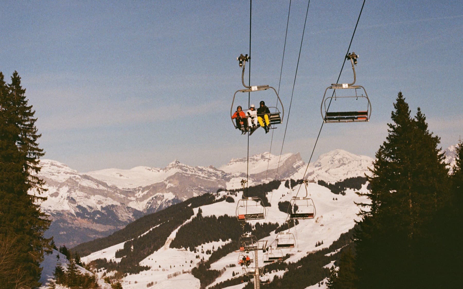 Chairlift carrying skiers uphill framed by dark pine trees, with snow-covered alpine peaks and a clear blue sky behind