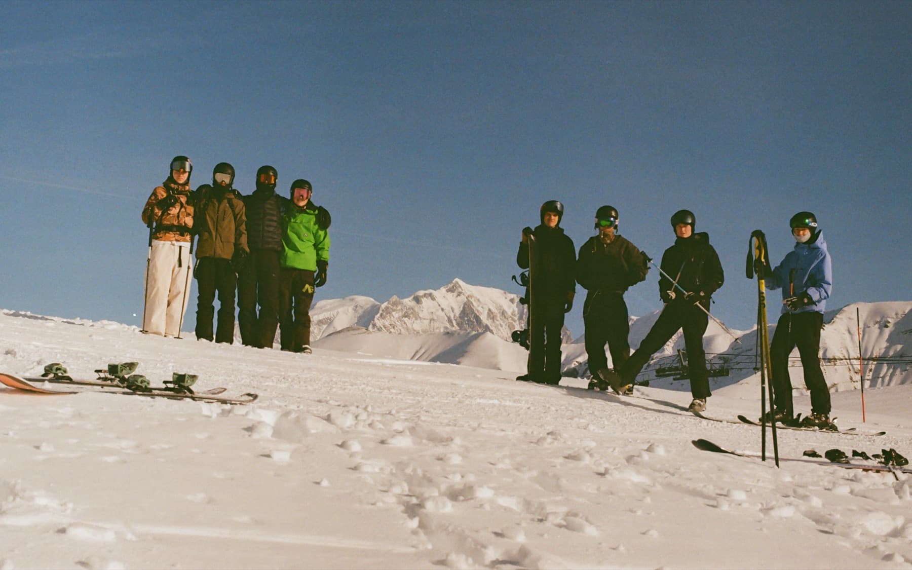 Group of eight skiers and snowboarders posing together on a snowy summit with a distant peak rising behind them under a clear blue sky