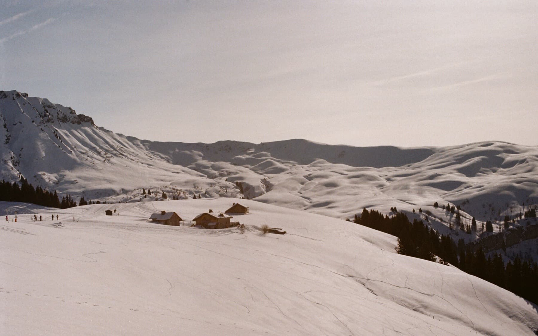 Remote alpine chalets nestled in a wide, untouched snow-covered valley with rolling white hills and a rocky ridge to the left